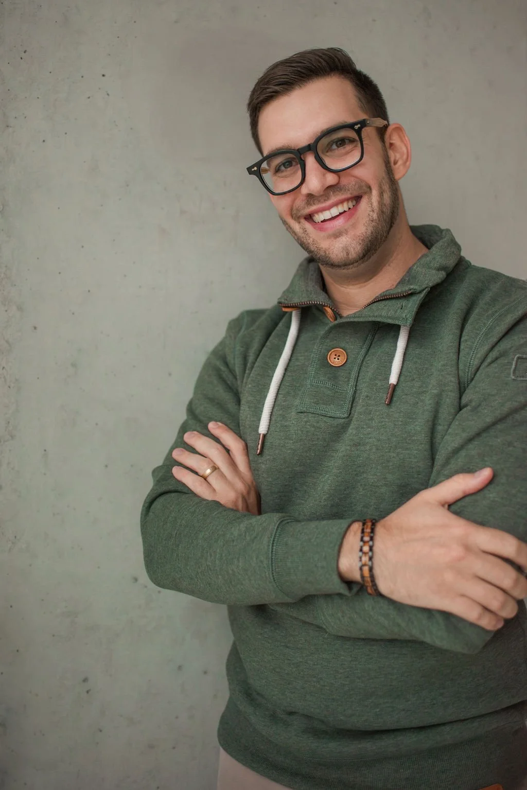 Ein lachender junger Mann mit Brille, grauem Hoodie und Armband, vor einer glatten, grauen Wand.