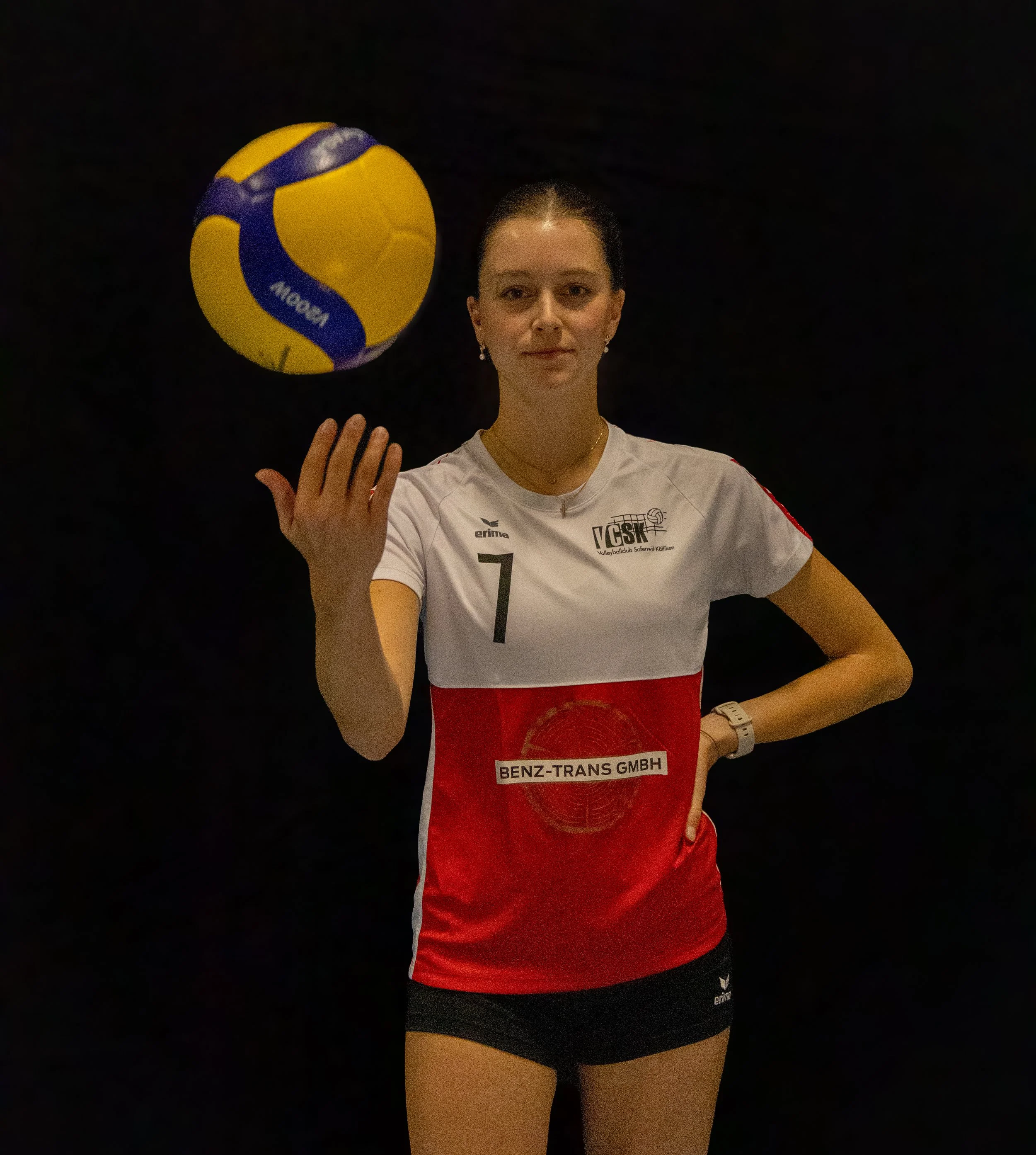 Junge Volleyballspielerin in weißem und rotem Sportoutfit auf einem schwarzen Hintergrund, die eine Volleyball ins Gesicht hält, lächelnd, mit Armbanduhr.