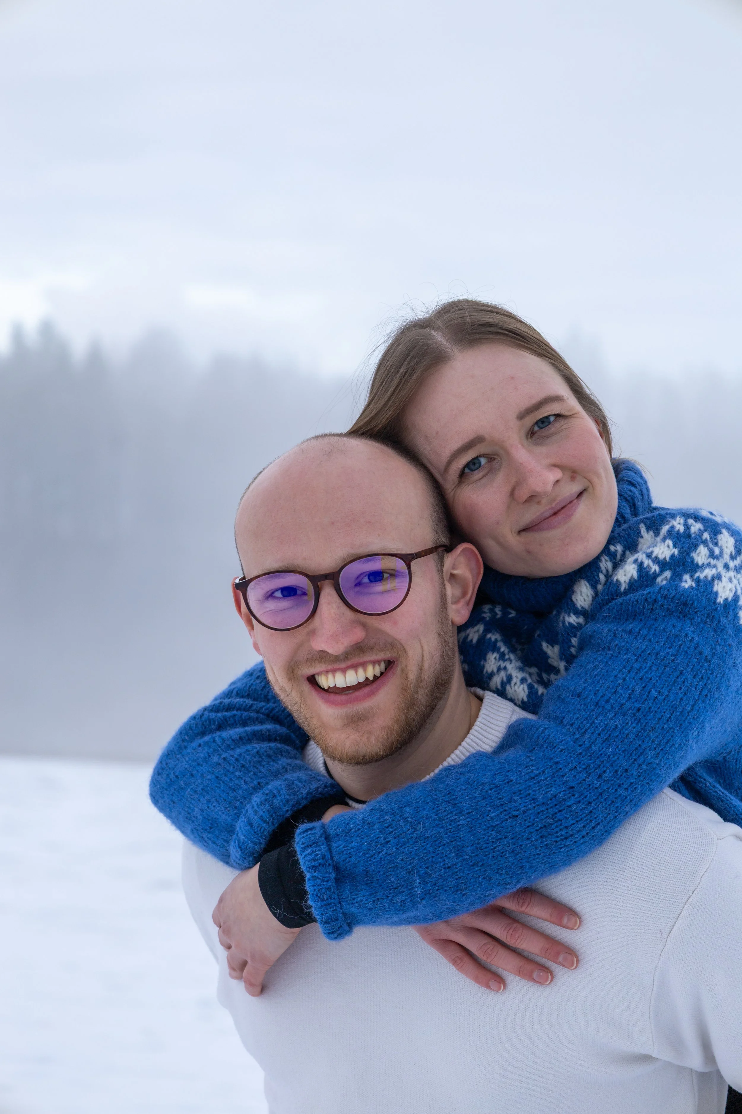 Zwei lachende Menschen, ein Mann mit Brille und ein lachendes Mädchen, die sich umarmen, im Schnee bei kaltem Wetter mit nebligem Hintergrund.