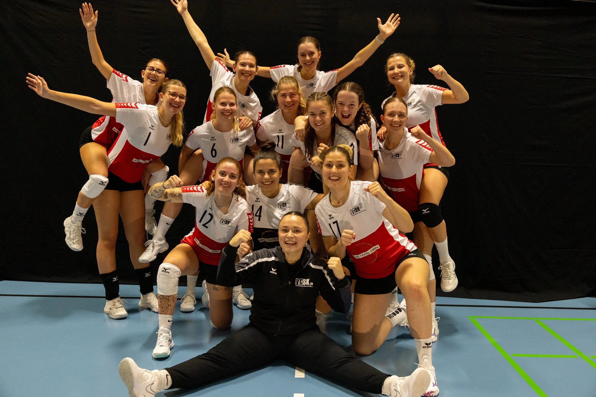 Eine Gruppe von weiblichen Volleyballspielerinnen in Sportuniformen, die jubeln und sich freuen, auf einem Hallenboden unter einem dunklen Hintergrund.