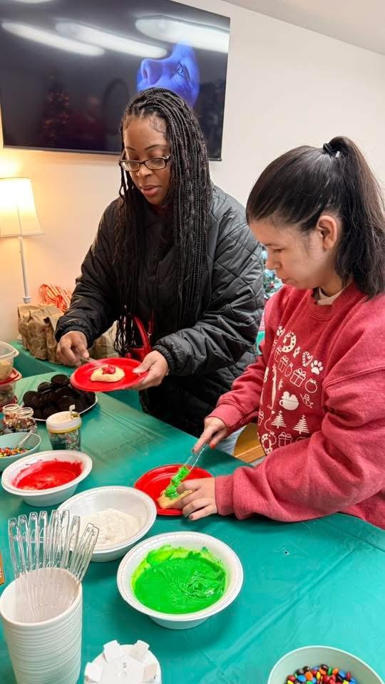 Two women decorating holiday cookies with green and red icing at a table with supplies, including bowls of colored icing, sprinkles, and cookies.
