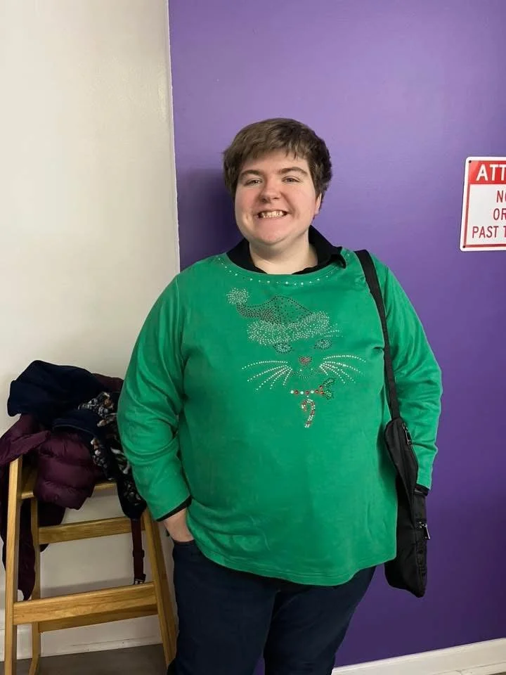 A person with short brown hair smiling while standing in front of a purple wall, wearing a green sweater with a Christmas-themed tiger design and carrying a black bag.