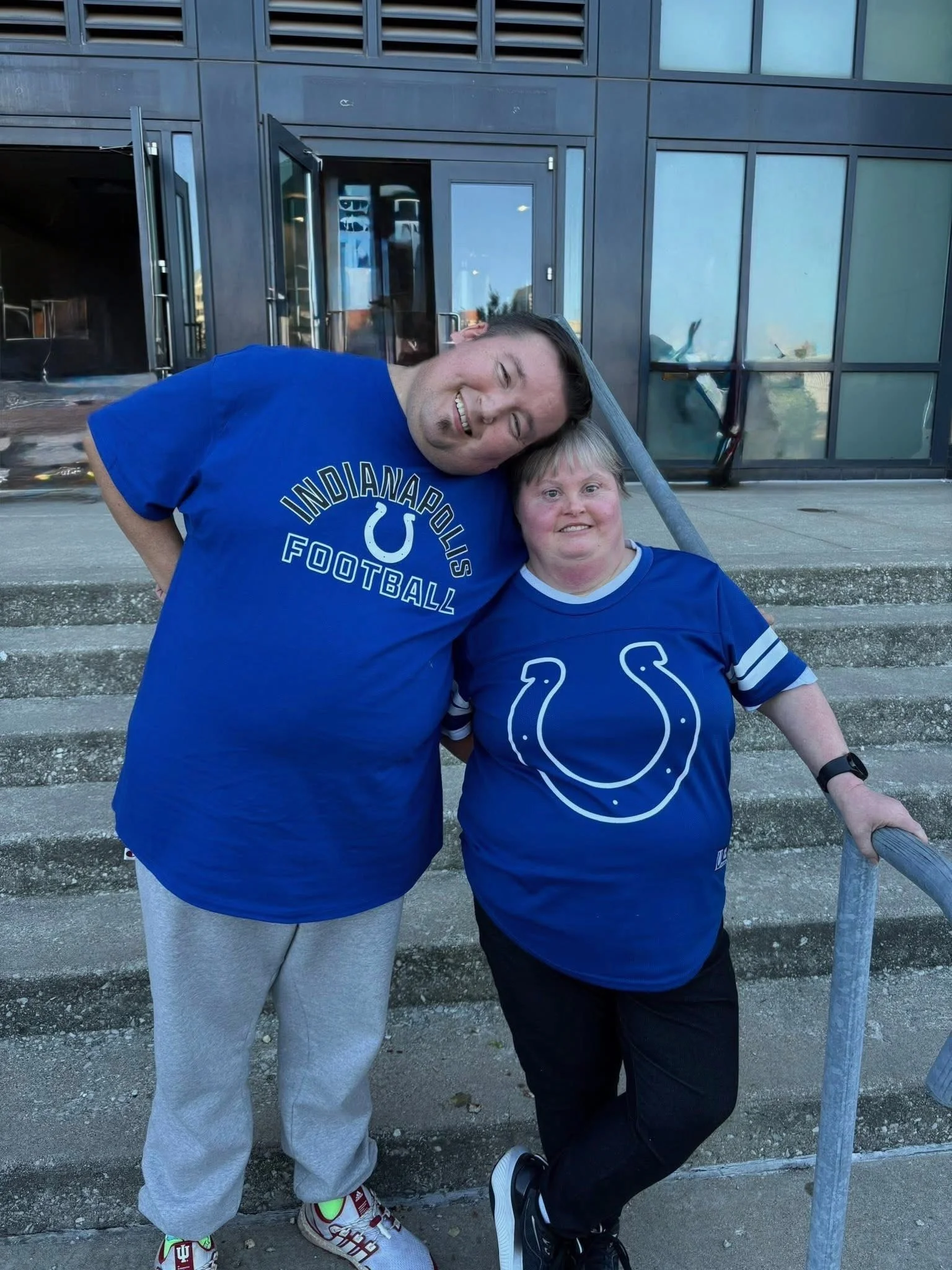 Two people standing on outdoor steps in front of a modern glass building, smiling and leaning their heads together, wearing blue Indianapolis Colts shirts.