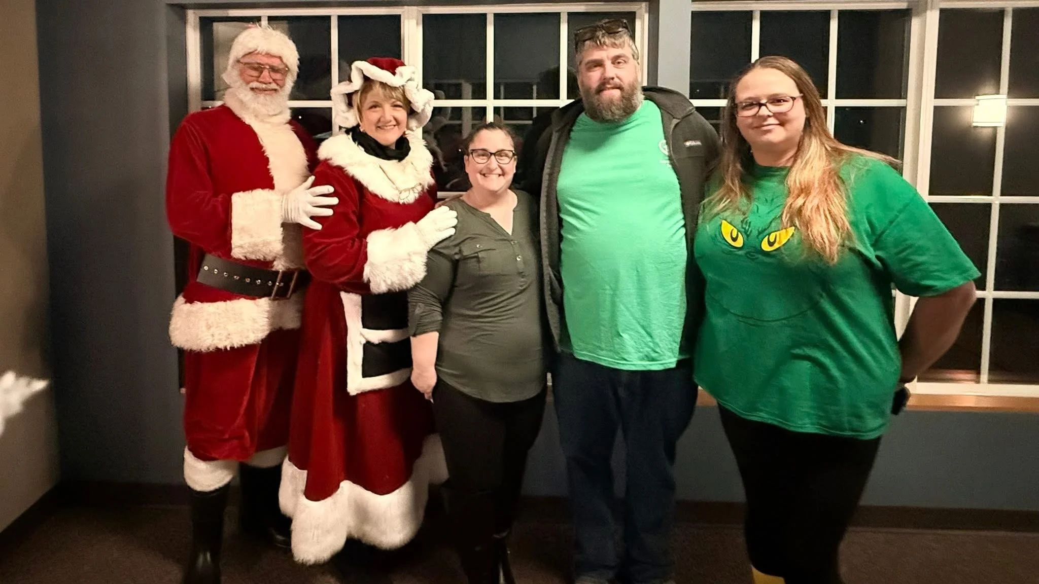 Five people standing together indoors at night, celebrating Christmas. Two dressed as Santa Claus and Mrs. Claus, the others in green shirts, one with a cat face design. They are smiling and posing in front of a large window with grid pattern.
