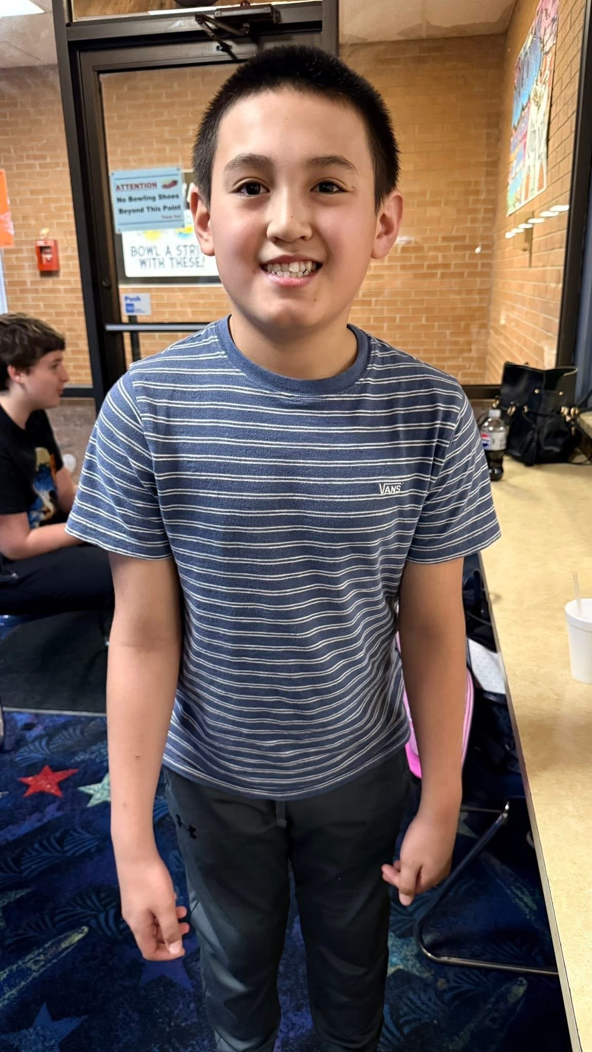 A smiling young boy with short black hair wearing a blue and white striped Vans t-shirt and dark pants, standing in a room with brick walls, next to a table with a white cup.