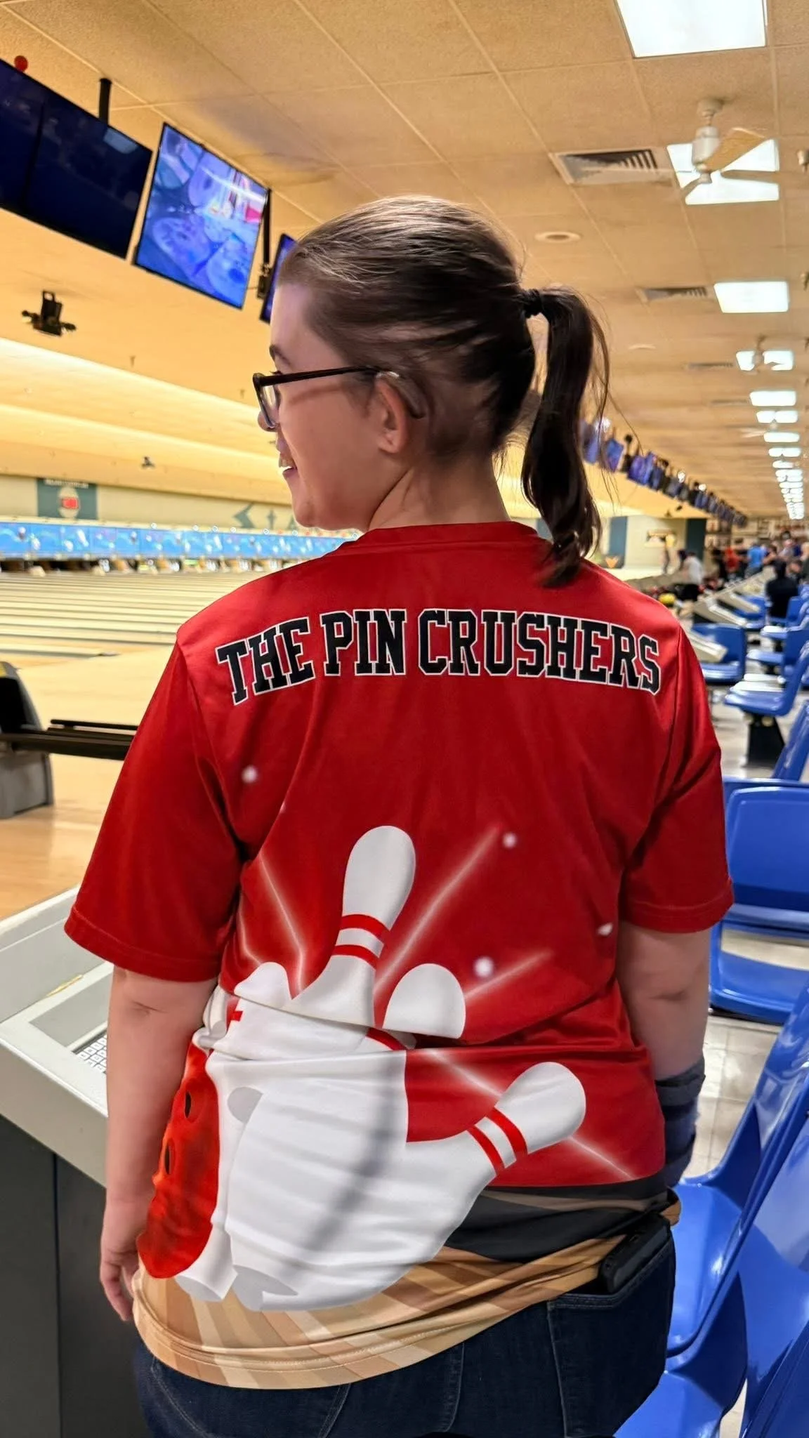 A woman with glasses in a red bowling shirt with the words "The Pin Crushers" and bowling pins graphic, standing in a bowling alley.