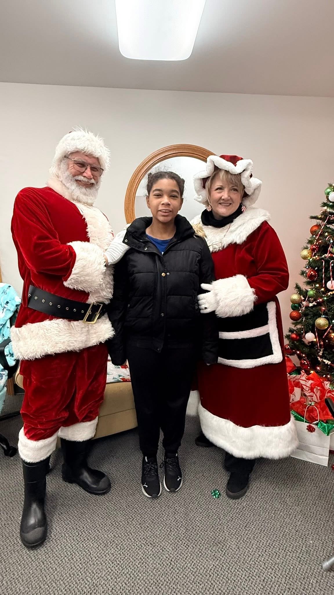 A young girl standing with Santa Claus and Mrs. Claus in front of a decorated Christmas tree indoors.