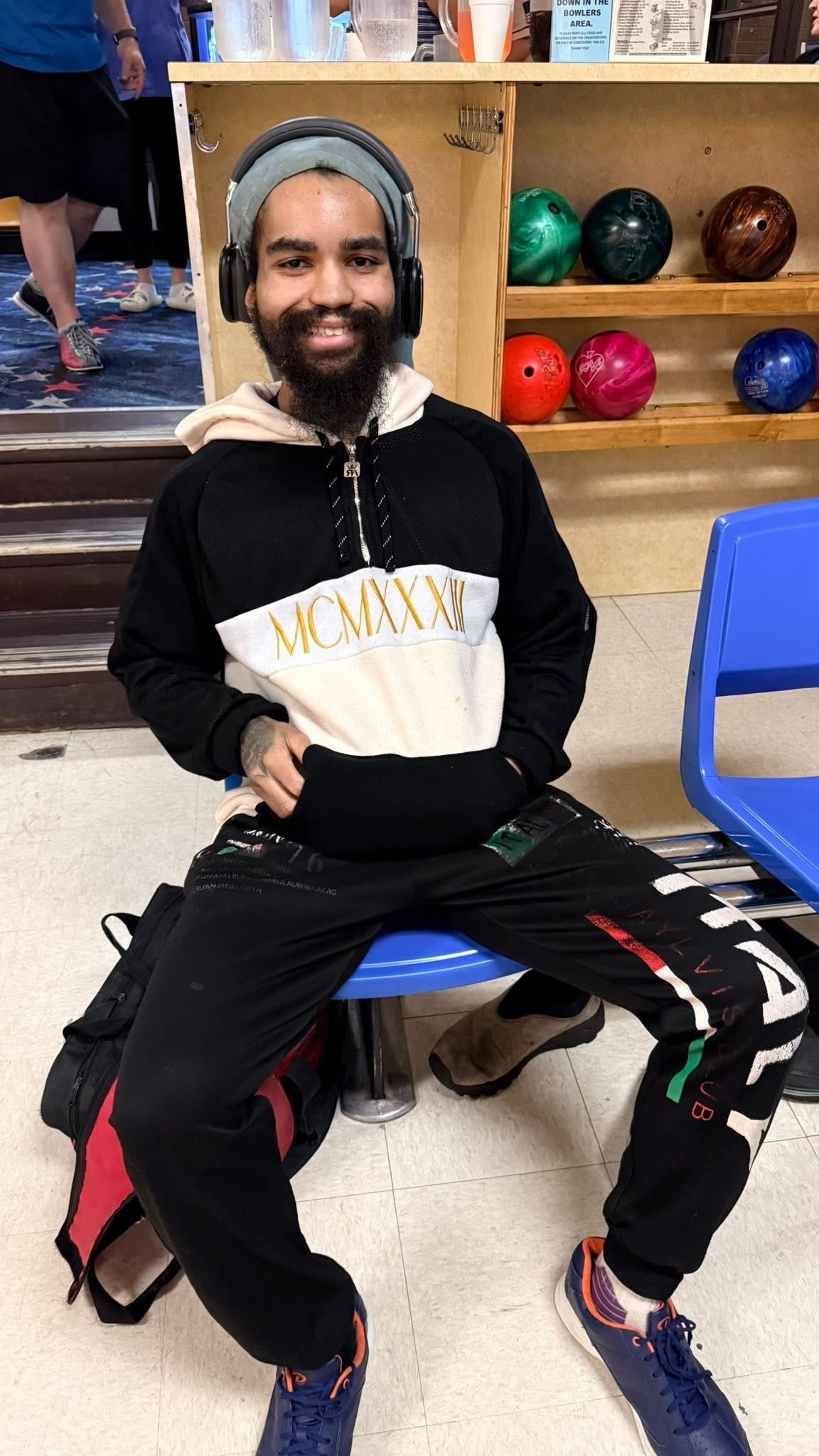 A man wearing headphones, a black and white sweatshirt with Roman numerals, black pants with branding, brown boots, and sitting on a blue bench at a bowling alley. There are colored bowling balls on shelves behind him.