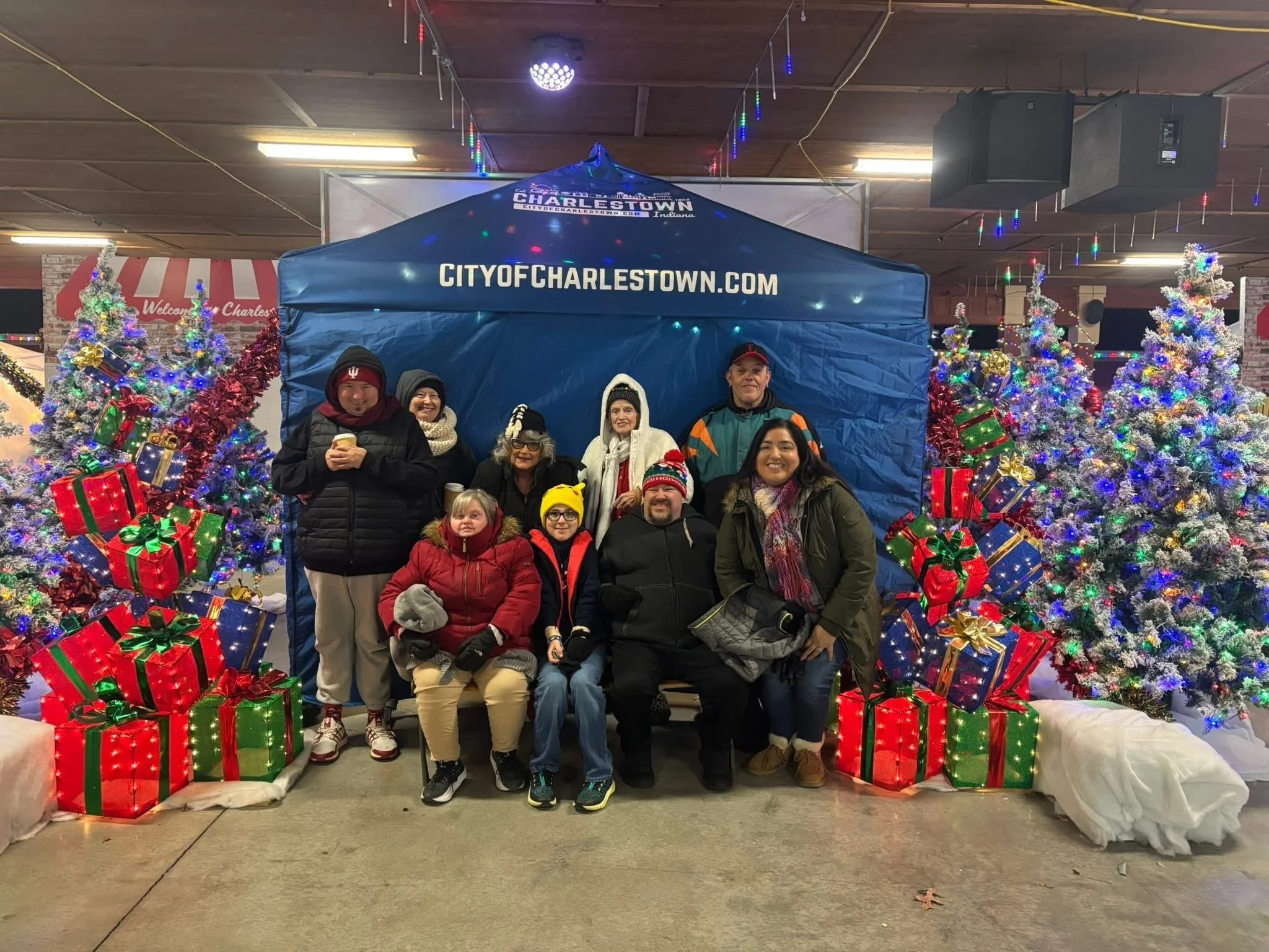 Group of people posing in front of Christmas trees decorated with lights and ornaments, surrounded by wrapped presents, at a festive holiday event in front of a blue canopy with a website URL and city name.