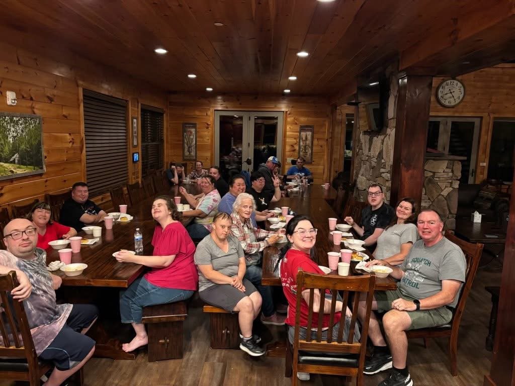 A group of people gathered around a long dining table in a wood-paneled room, enjoying a meal together with plates and drinks, smiling and looking at the camera.