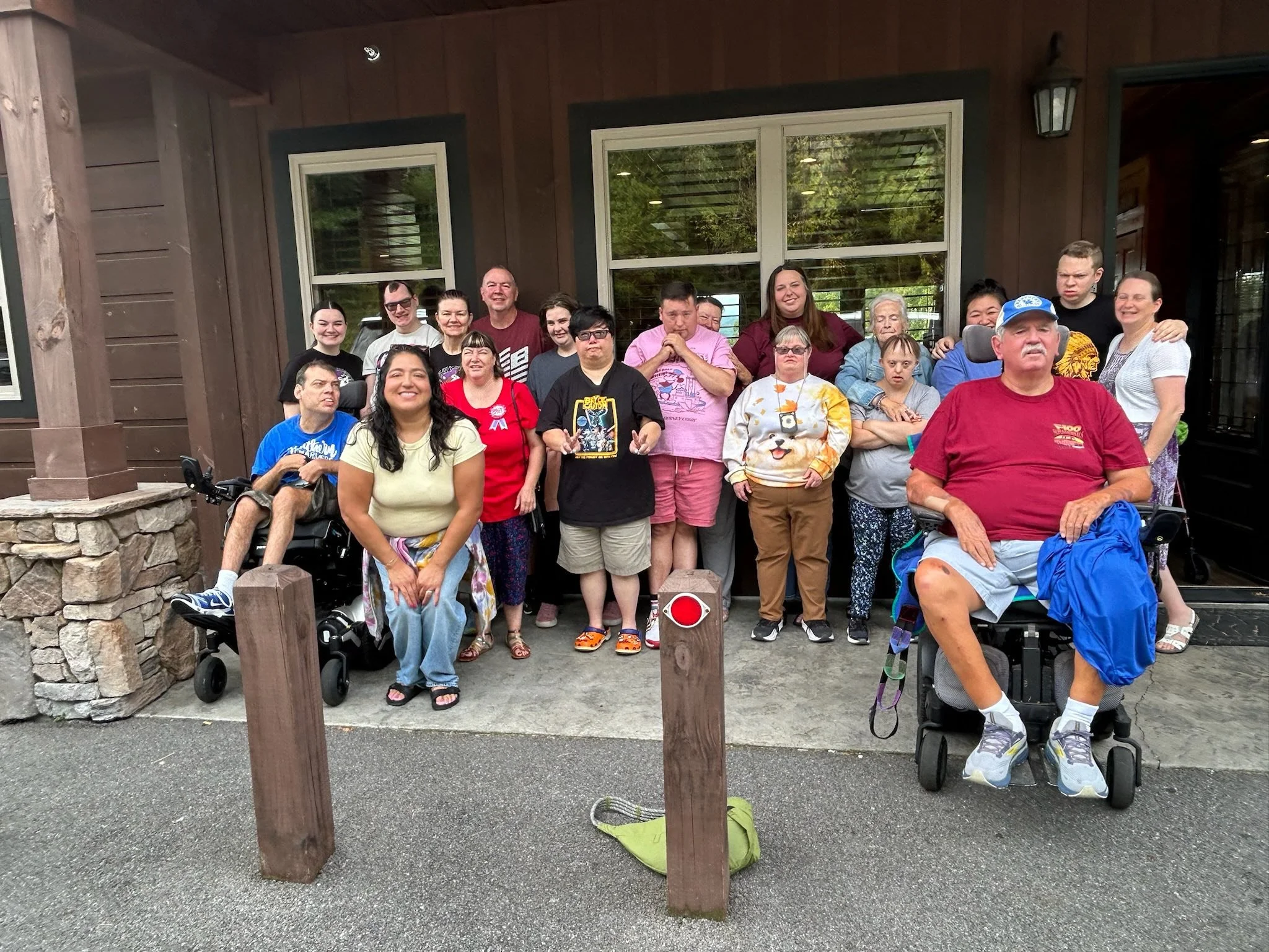Group of diverse people, some in wheelchairs, standing and sitting outside a building with wooden siding and large windows, posing for a group photo.