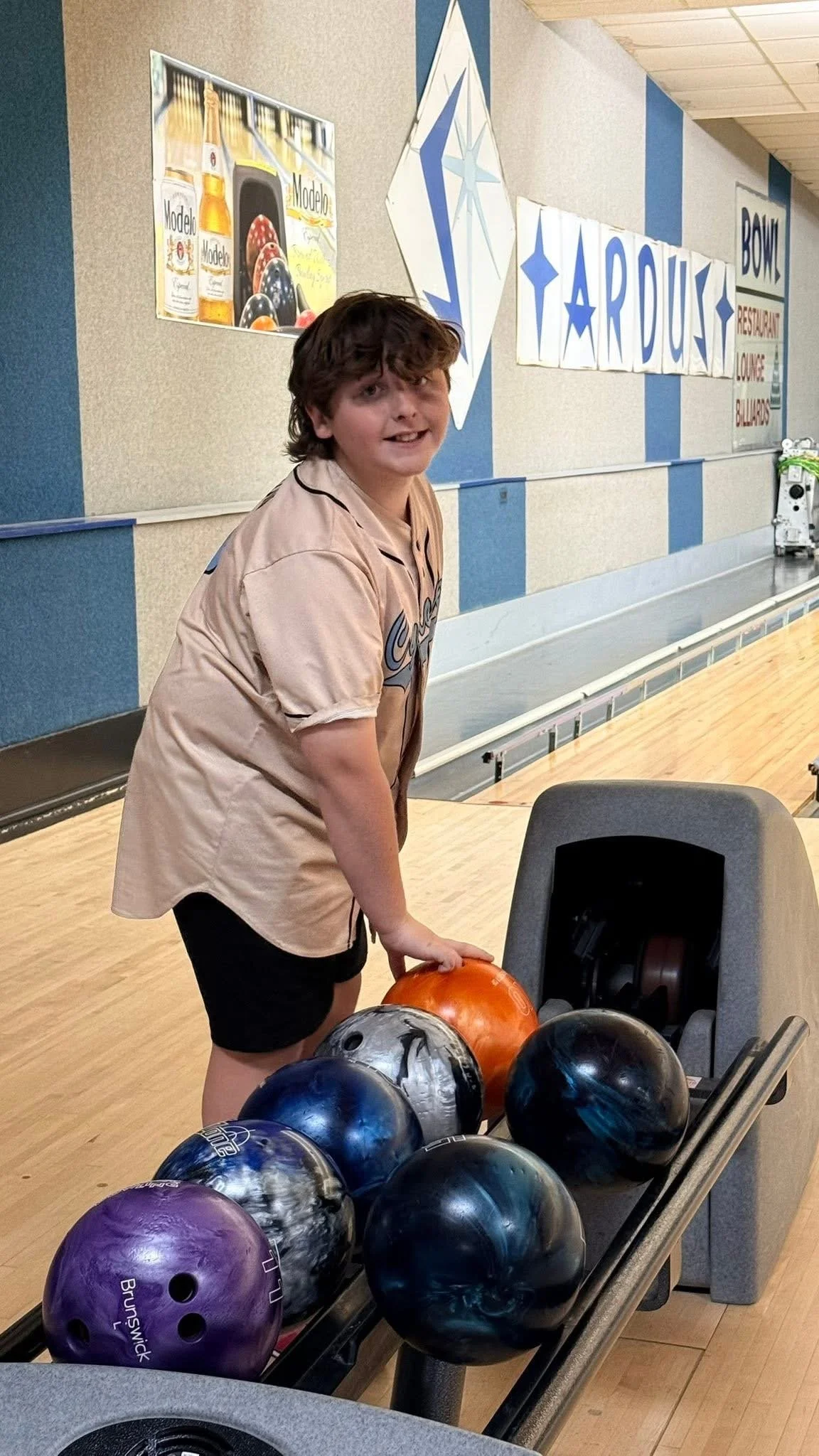 A young boy at a bowling alley placing his hand on an orange bowling ball, with several other balls on the ball return rack in front of him. The bowling alley has a blue and beige color scheme, and there are signs and advertisements on the wall behin