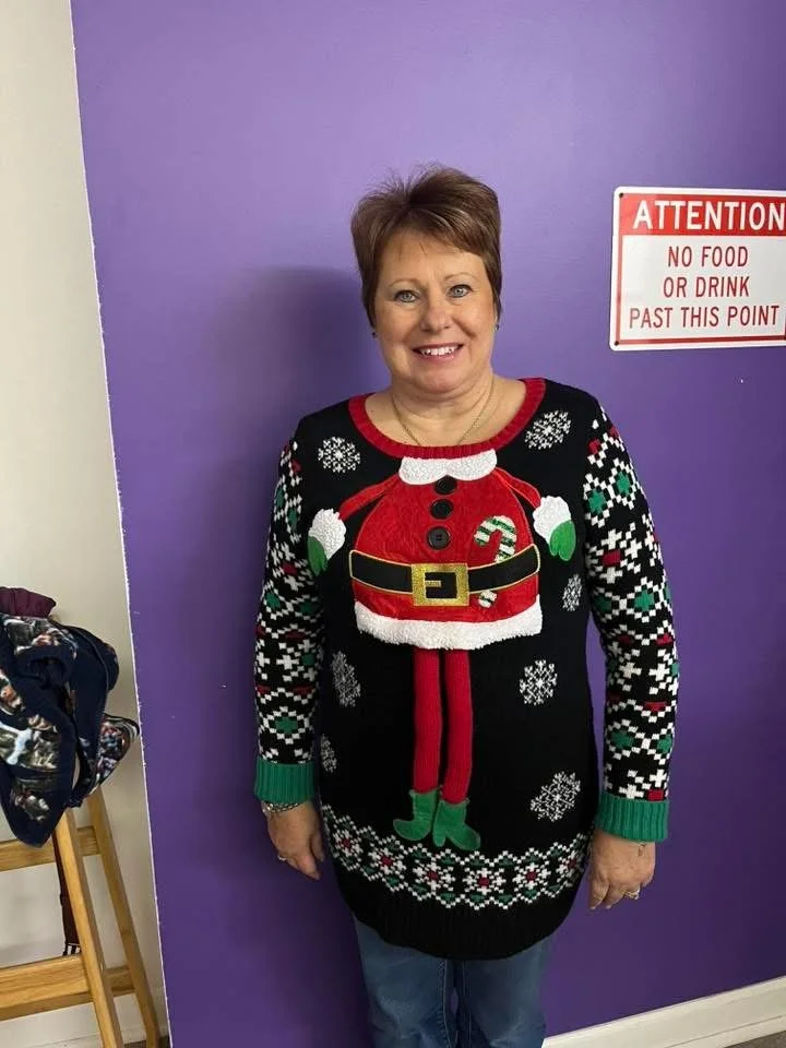 Woman smiling wearing a Christmas sweater with a Santa Claus design, standing in front of a purple wall with a no food or drink sign.