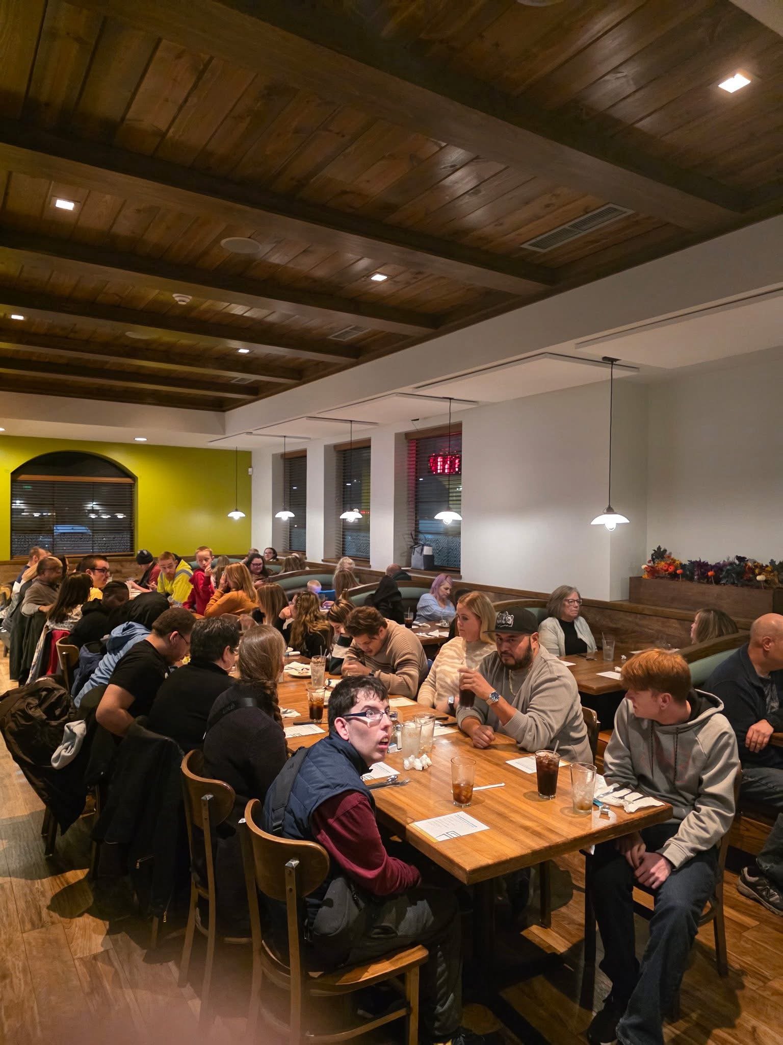 People dining at a long table in a restaurant with wood-paneled ceiling and modern lighting.