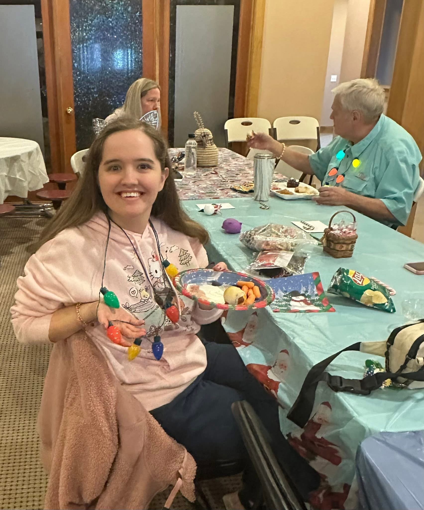 A girl at a Christmas party holding a plate of snacks, sitting at a decorated table with others in the background.
