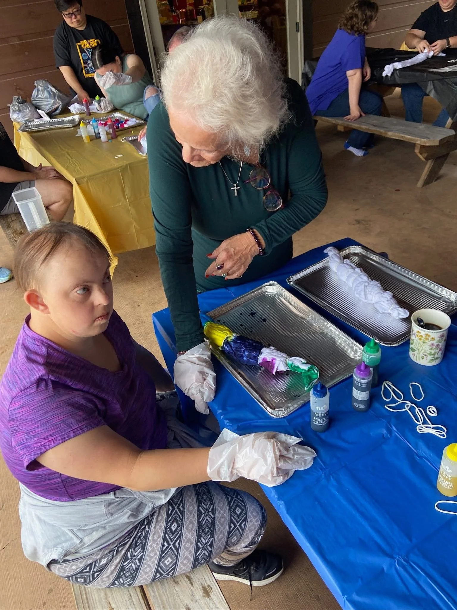 An elderly woman and a young girl wearing gloves sit at a table with art supplies, creating tie-dye designs on fabric. Several trays with tied-dye items are on the table, and other people in the background are engaged in similar activities.