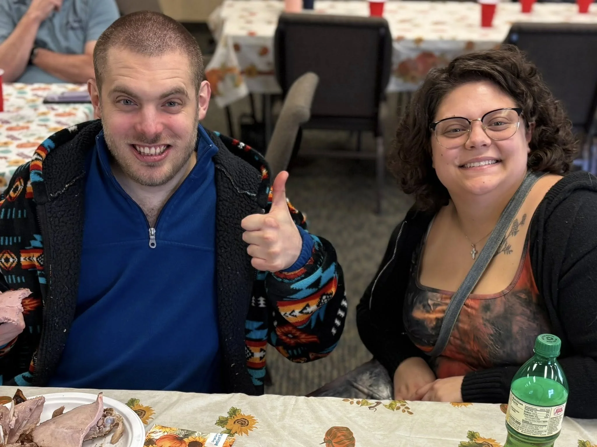 A man and woman sitting at a table, smiling and posing for the camera. The man is giving a thumbs up, wearing a blue shirt and a colorful jacket. The woman has curly hair, glasses, and a tattoo on her chest, wearing a print top and black jacket. Ther