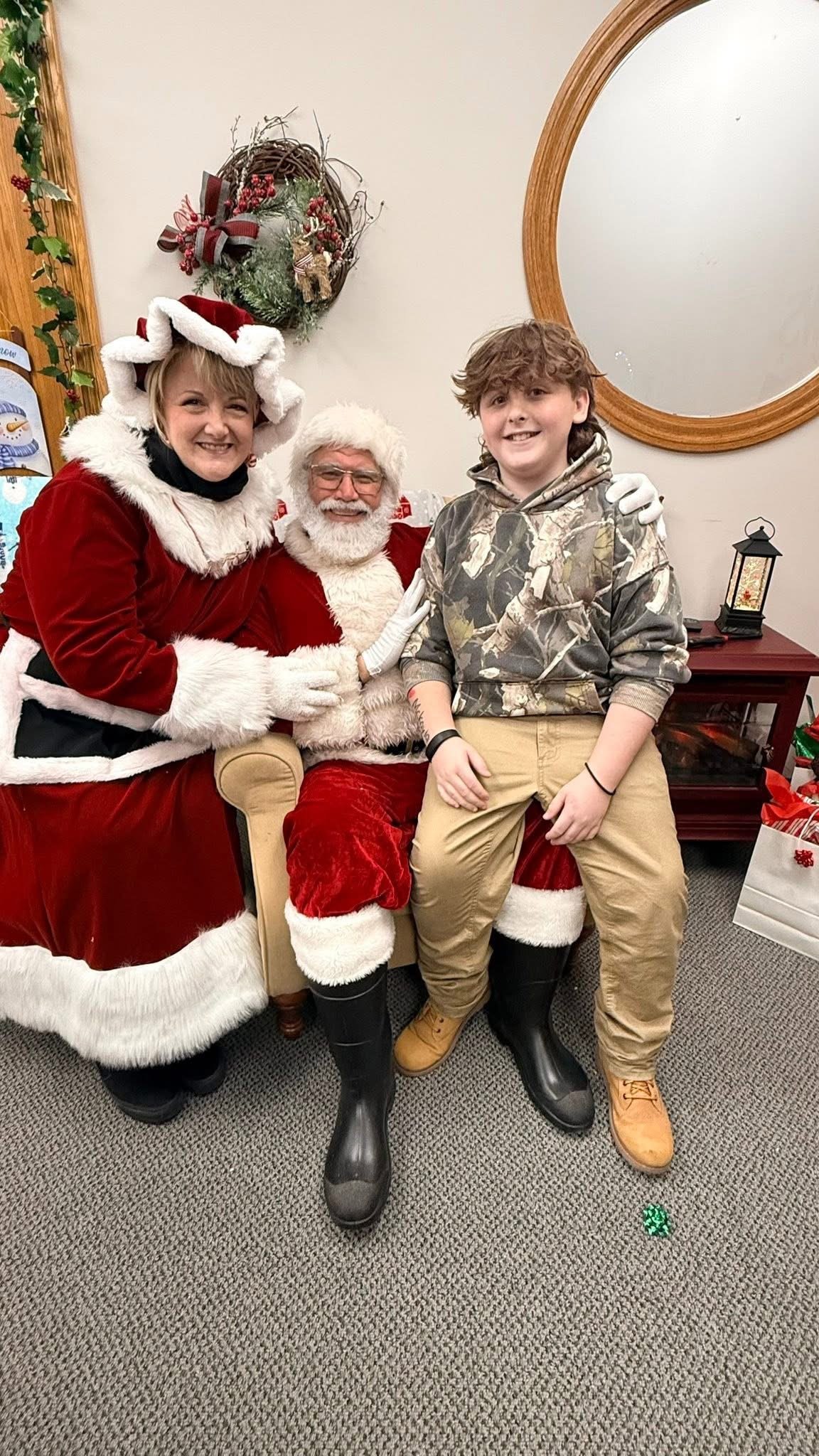 A young man sitting on Santa Claus's lap, both smiling, with a woman dressed as Mrs. Claus standing next to Santa, all in a festive setting with Christmas decorations.