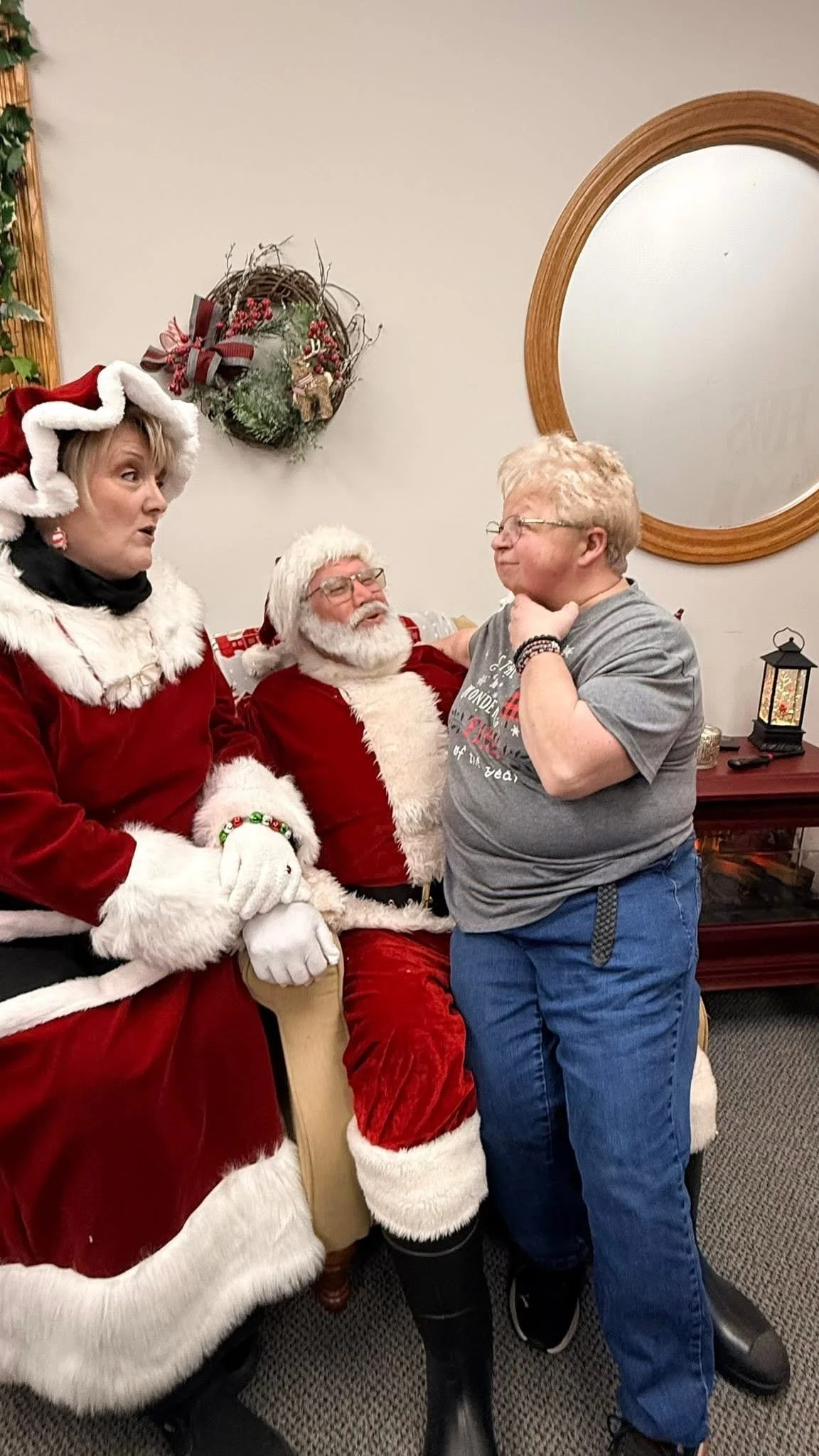 A woman dressed as Mrs. Claus, a man dressed as Santa Claus, and a woman are having a conversation indoors. Santa is seated, while Mrs. Claus and the woman stand near him. The woman standing has her hand on her chin and appears to be talking or expla