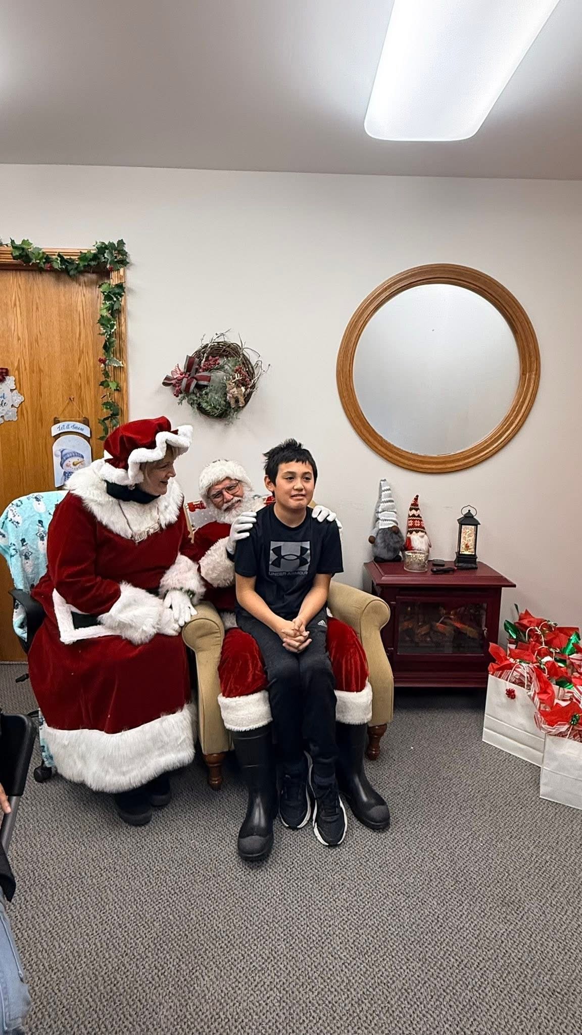 A young boy sitting on Santa's lap, with Mrs. Claus standing beside them, posing for a holiday photo inside a decorated room with Christmas ornaments and gifts.