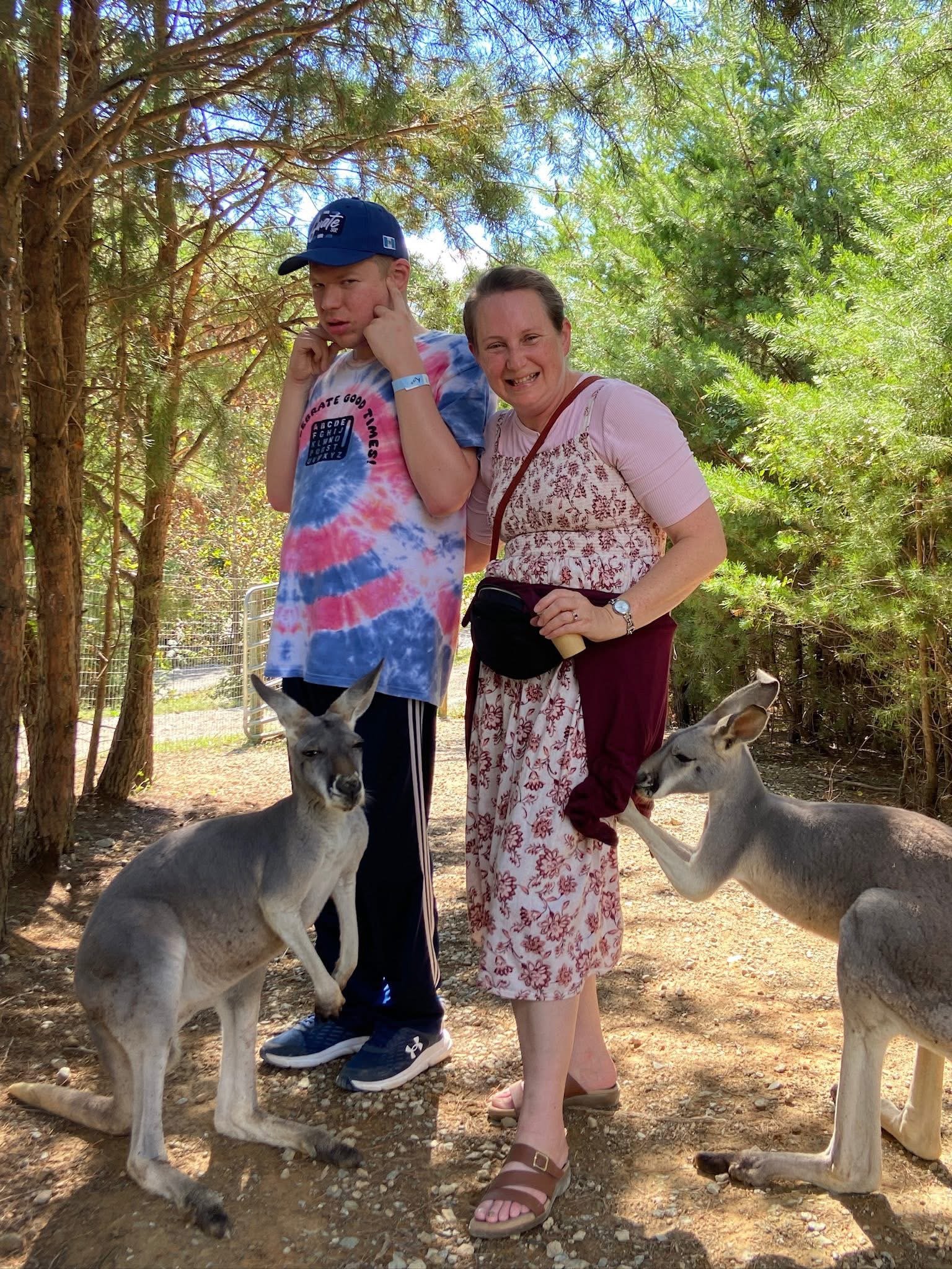 A woman and a boy are standing in a wooded area with trees and sunlight. The boy is wearing a tie-dye shirt, a blue cap, and dark pants, and they are touching their faces. The woman is smiling, wearing a pink top with a floral pattern, a maroon skirt