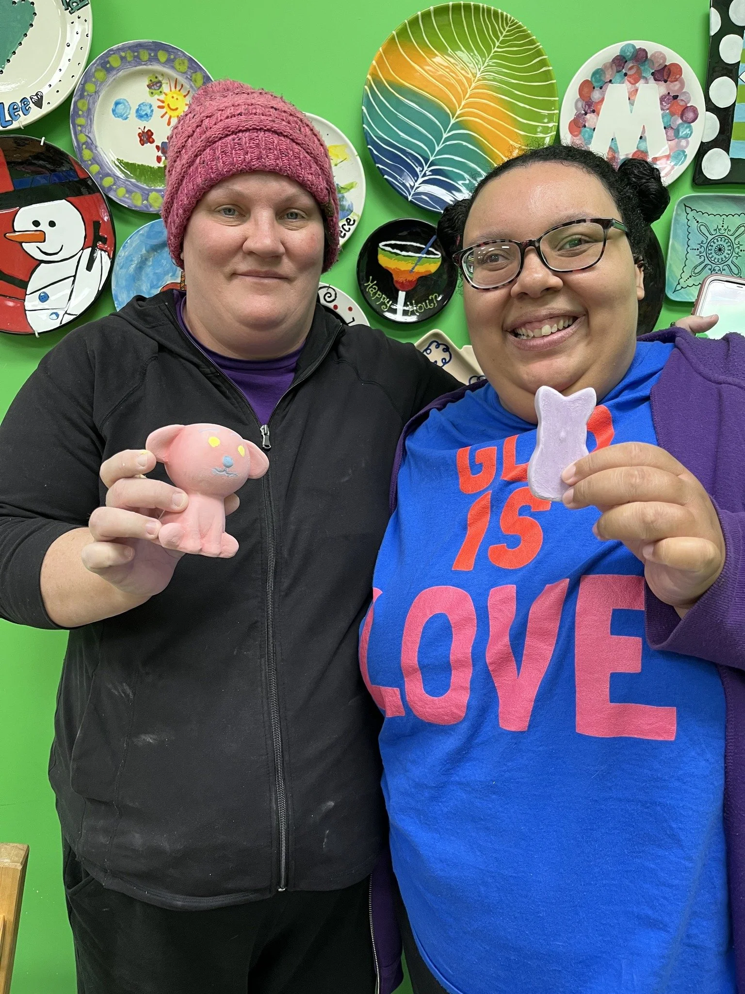 Two women smiling and holding handmade clay figurines, standing in front of a colorful wall decorated with various painted plates.