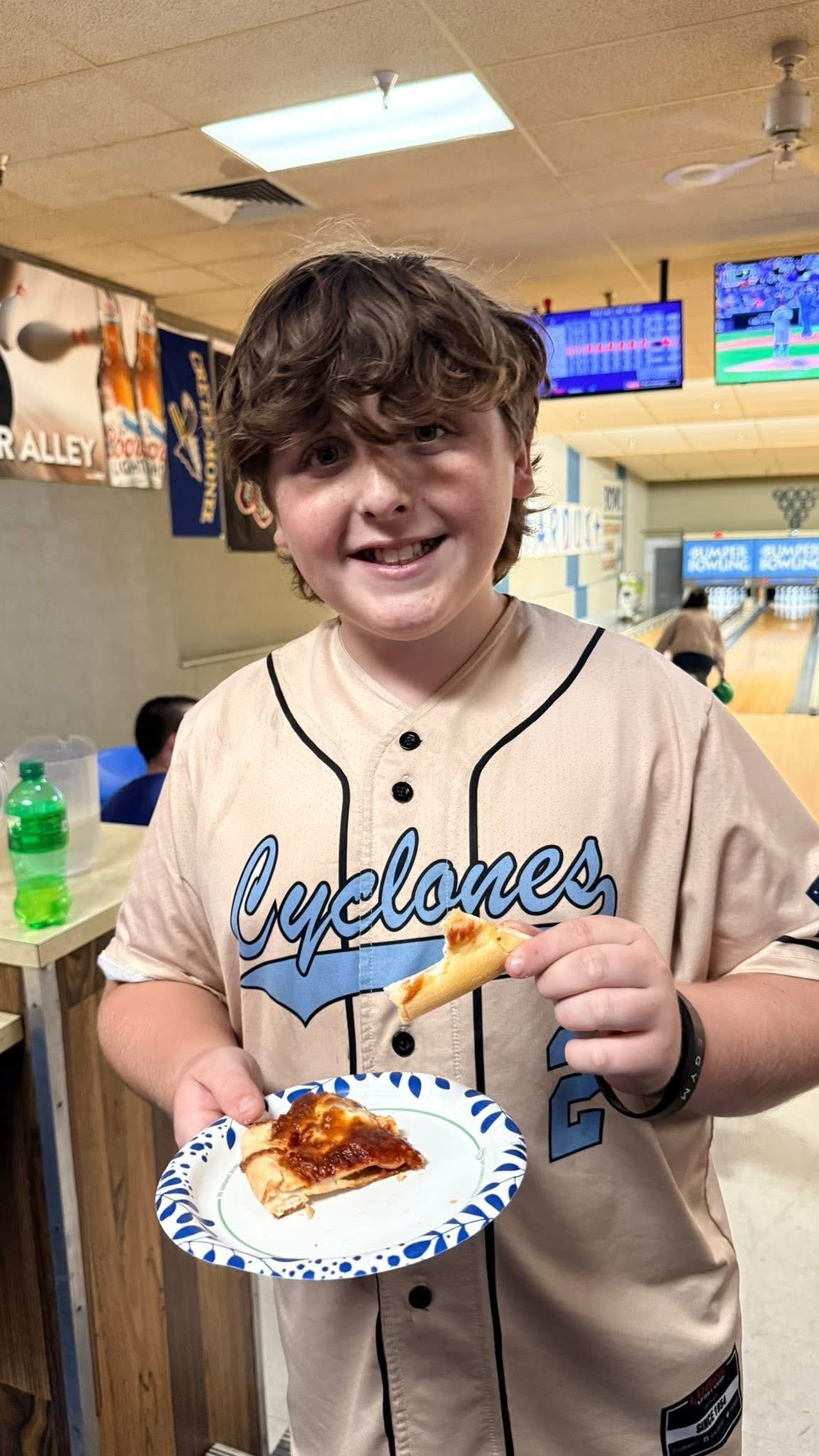 A young boy at a bowling alley holding a small pizza and slice of pizza, smiling at the camera.