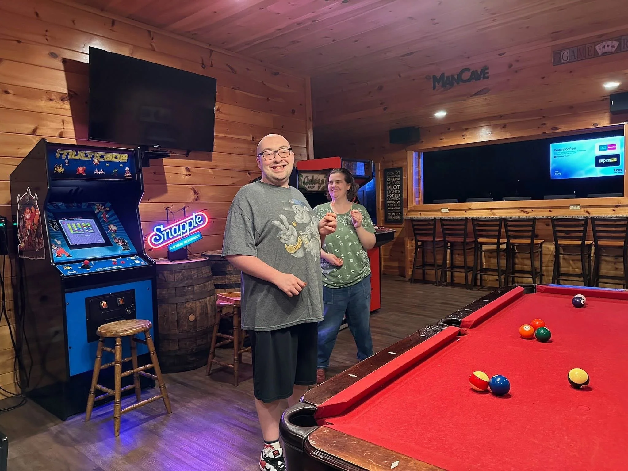 Two people playing pool in a wooden bar, with one person holding a pool cue and the other smiling. The room has arcade games, a large TV, and bar stools.