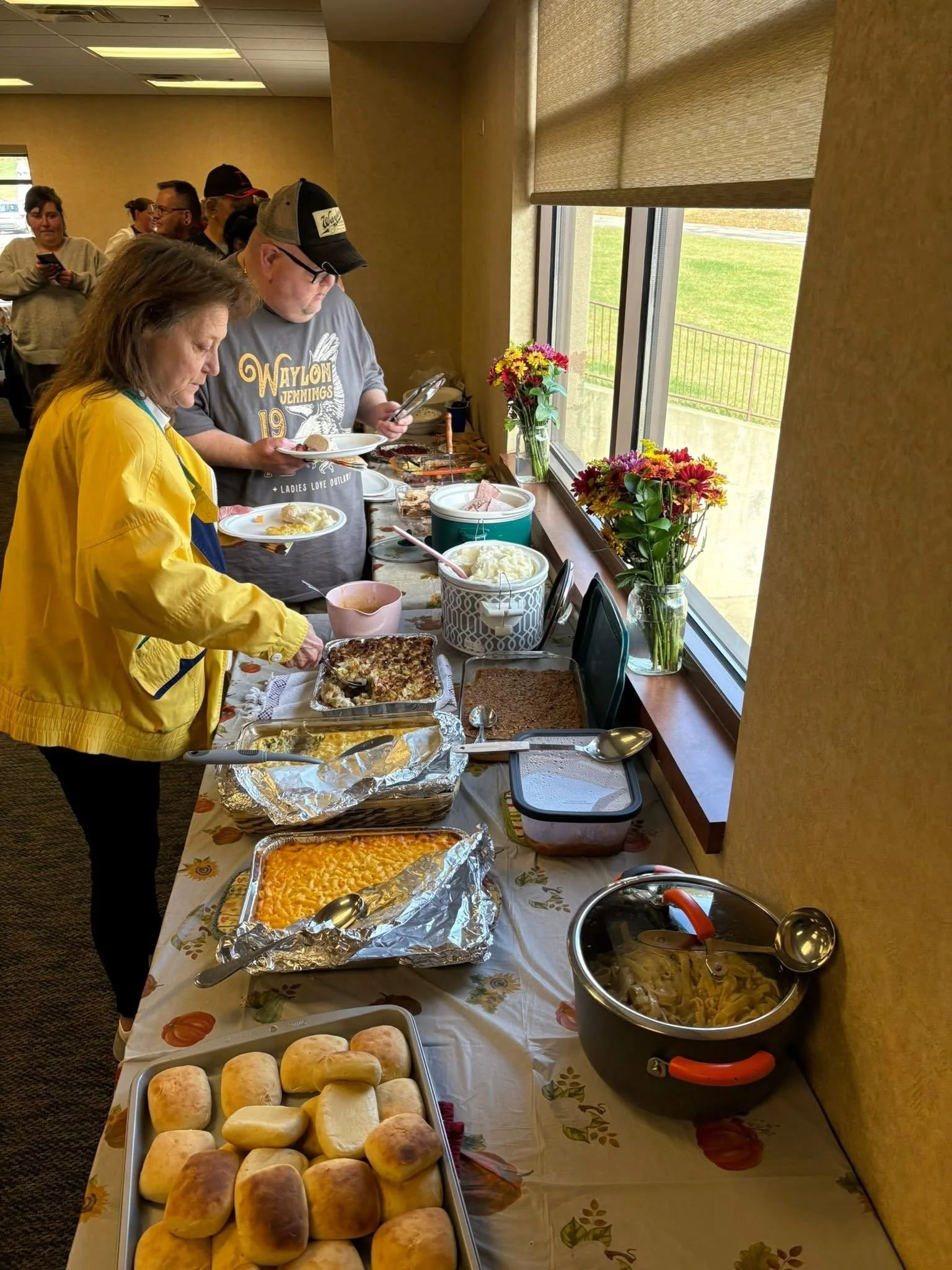 People serving themselves food at a buffet table with baked bread rolls, casseroles, salads, and pasta, inside a room with large windows and flower arrangements.
