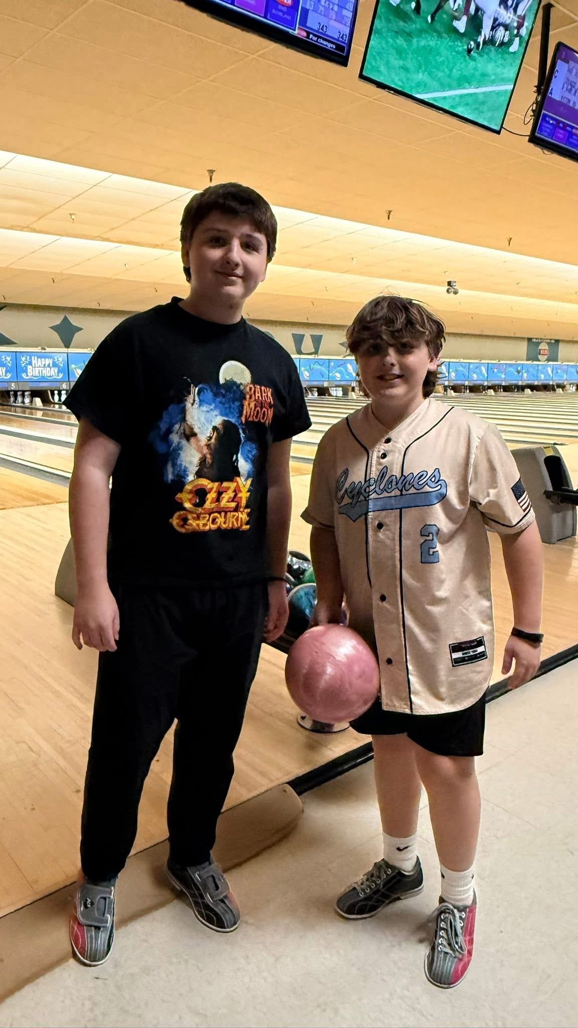 Two boys standing in a bowling alley, one of them holding a pink bowling ball.