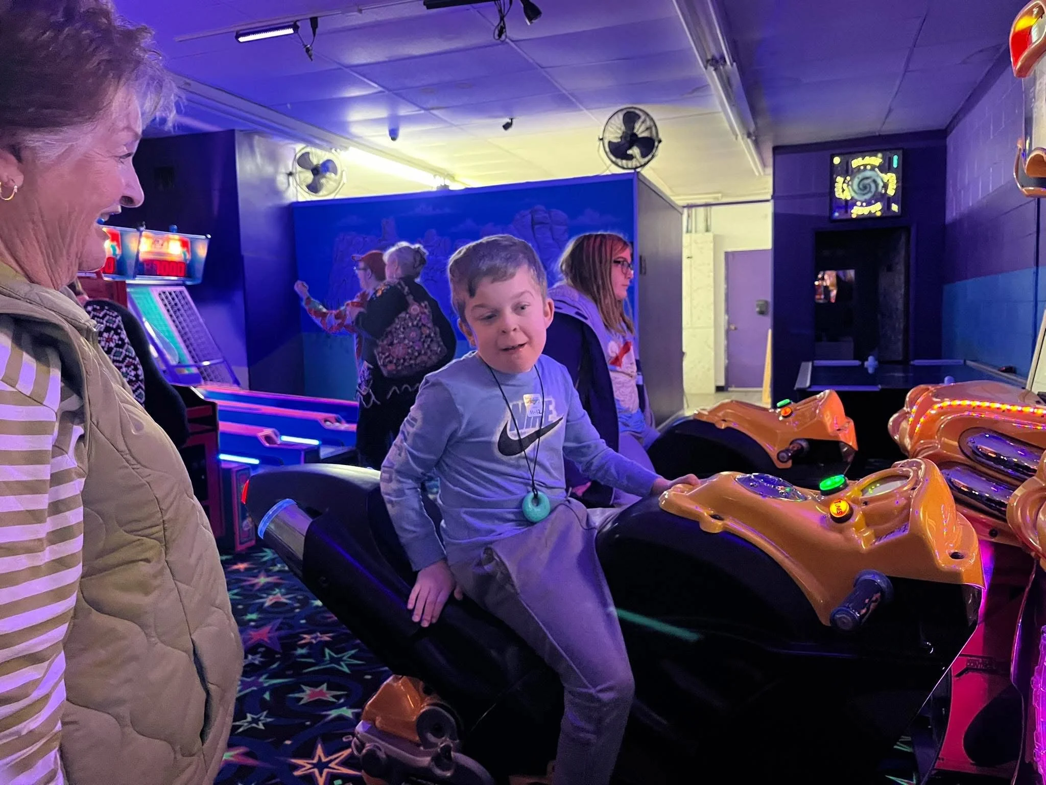 A young boy on an arcade motorcycle ride looks surprised, with a woman in a beige vest and striped shirt watching him, surrounded by other children and arcade machines in a colorful, neon-lit arcade.
