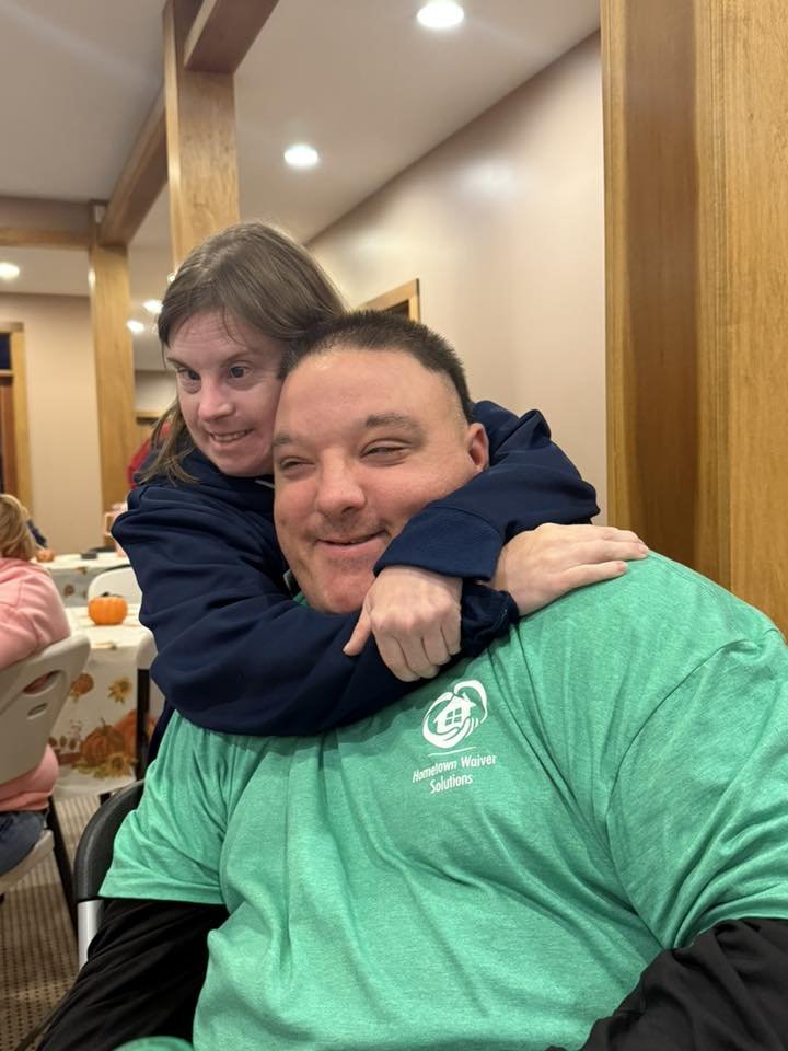 A young girl with long brown hair hugging a man with short dark hair, both smiling and appearing happy, in a warmly lit indoor setting.