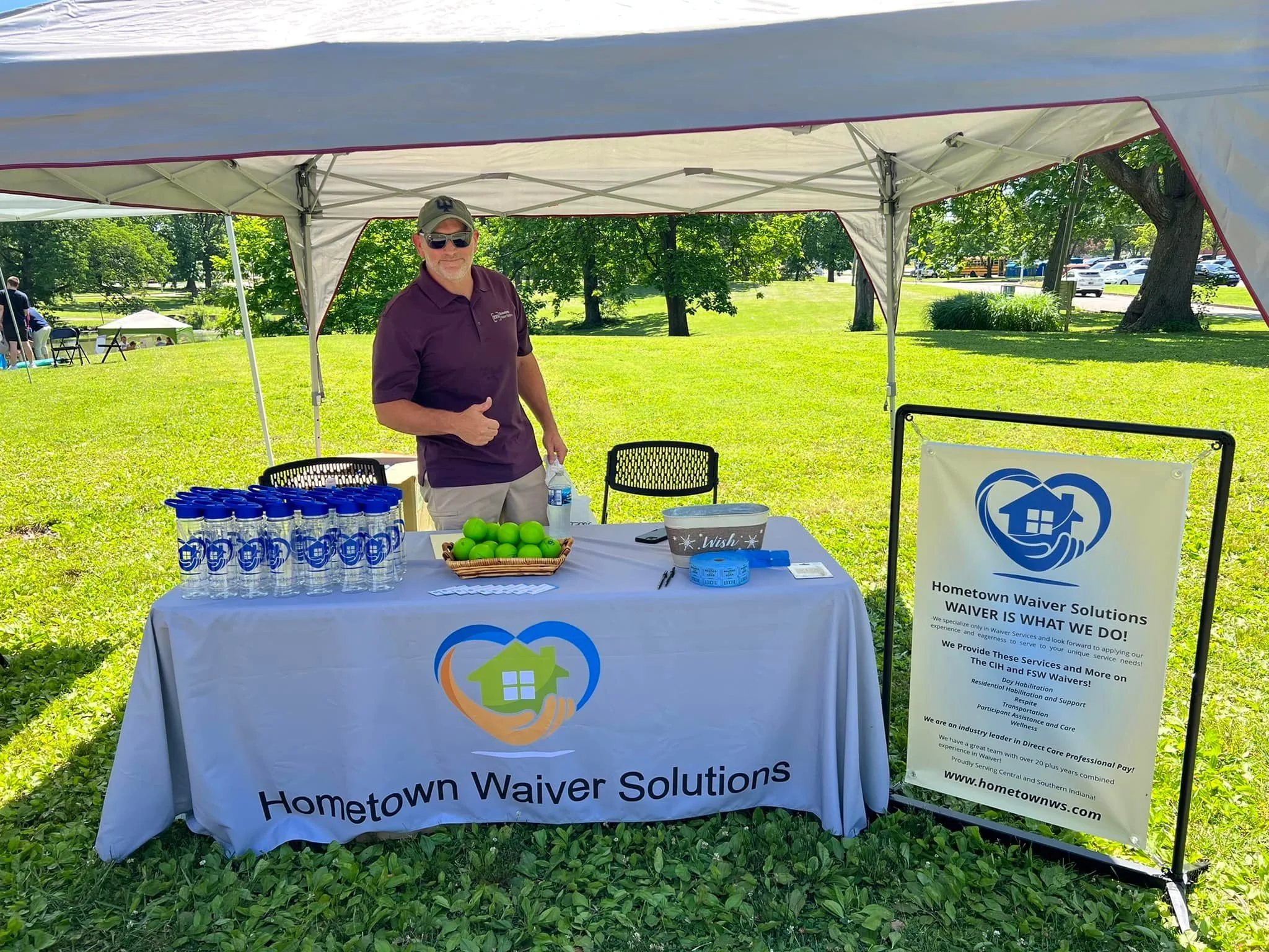 A man standing behind a table under a tent at an outdoor event. The table has a banner with the logo and name 'Hometown Waiver Solutions,' along with bottled water, green tennis balls, and writing materials. A large sign next to the table displays in