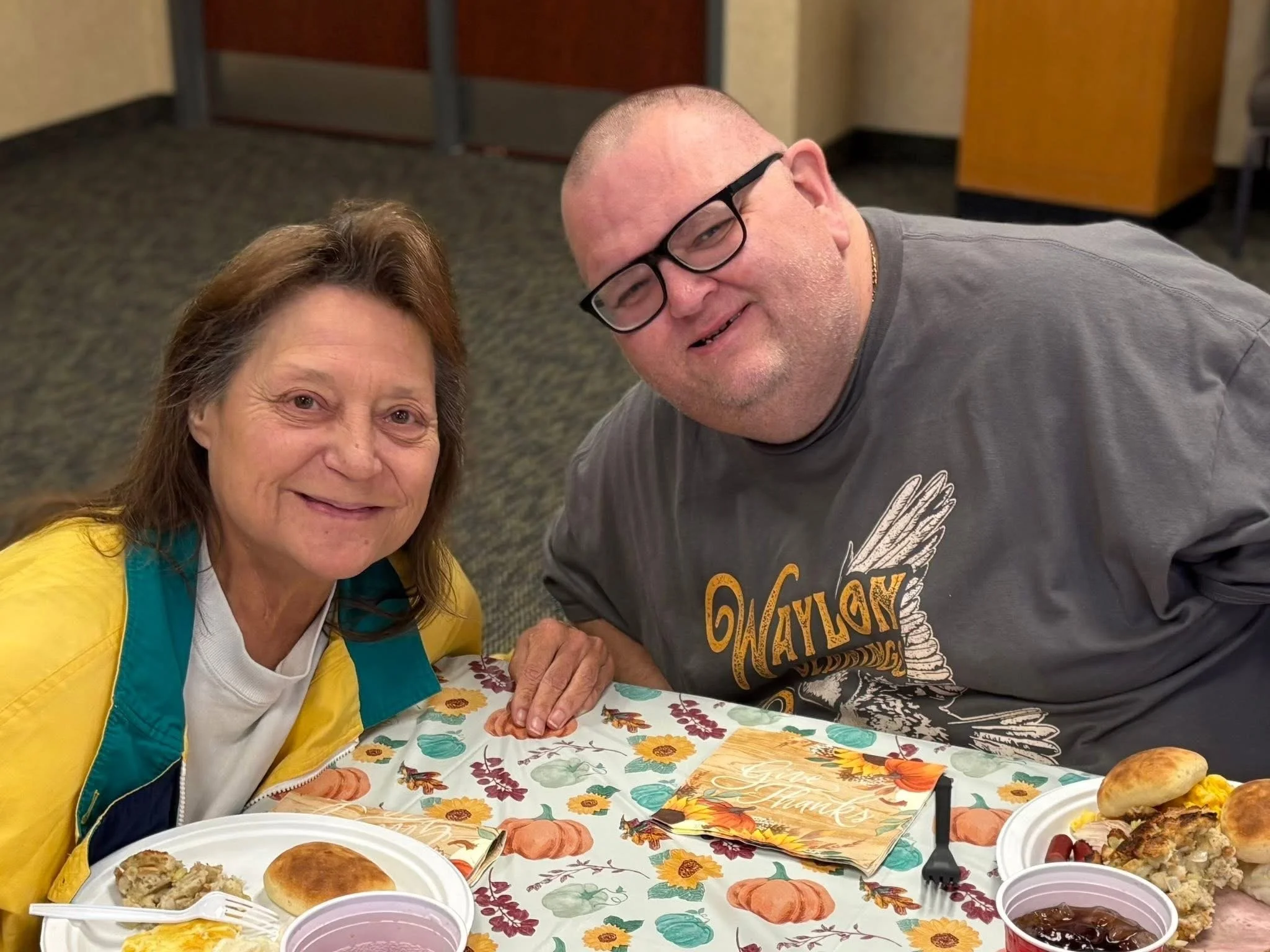 A smiling woman and man sitting at a table with fall-themed tablecloth, food, and a thank you card, celebrating Thanksgiving.