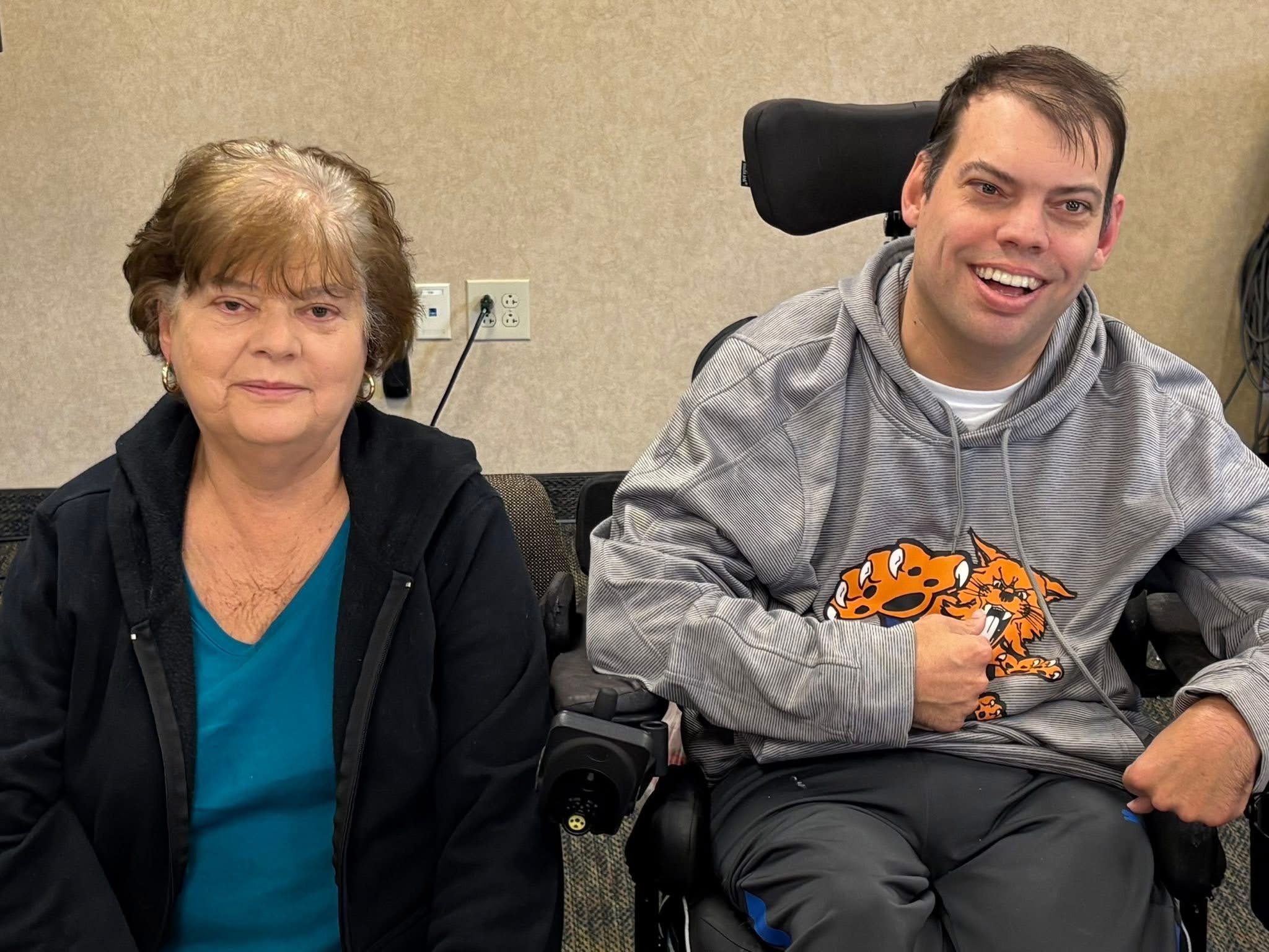 An elderly woman and a young man smiling at the camera. The young man is in a motorized wheelchair and wearing a gray hoodie with an orange graphic of a wildcat.