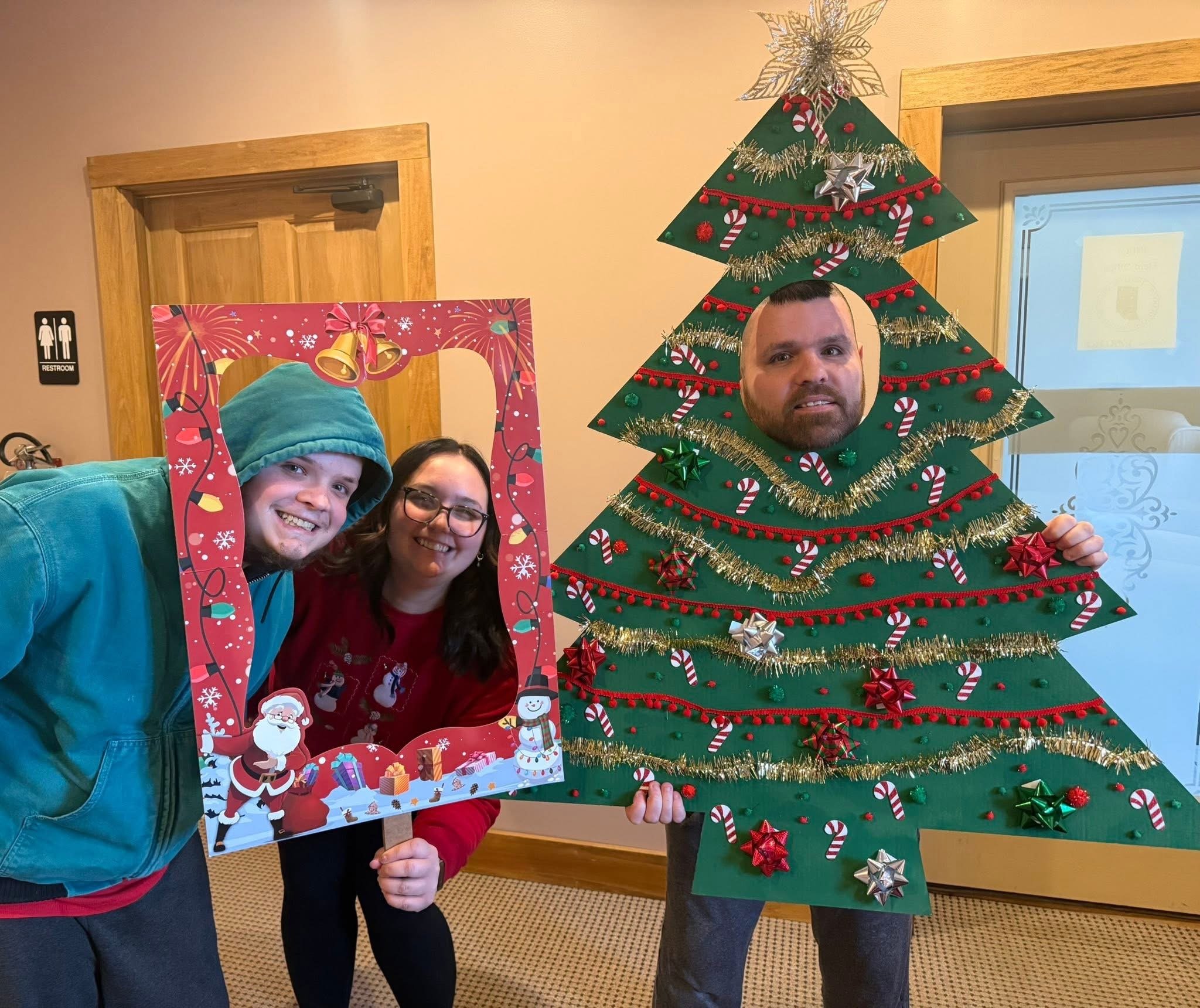 People celebrating Christmas with a tree and festive decorations, one person holding a Christmas-themed photo frame, others nearby