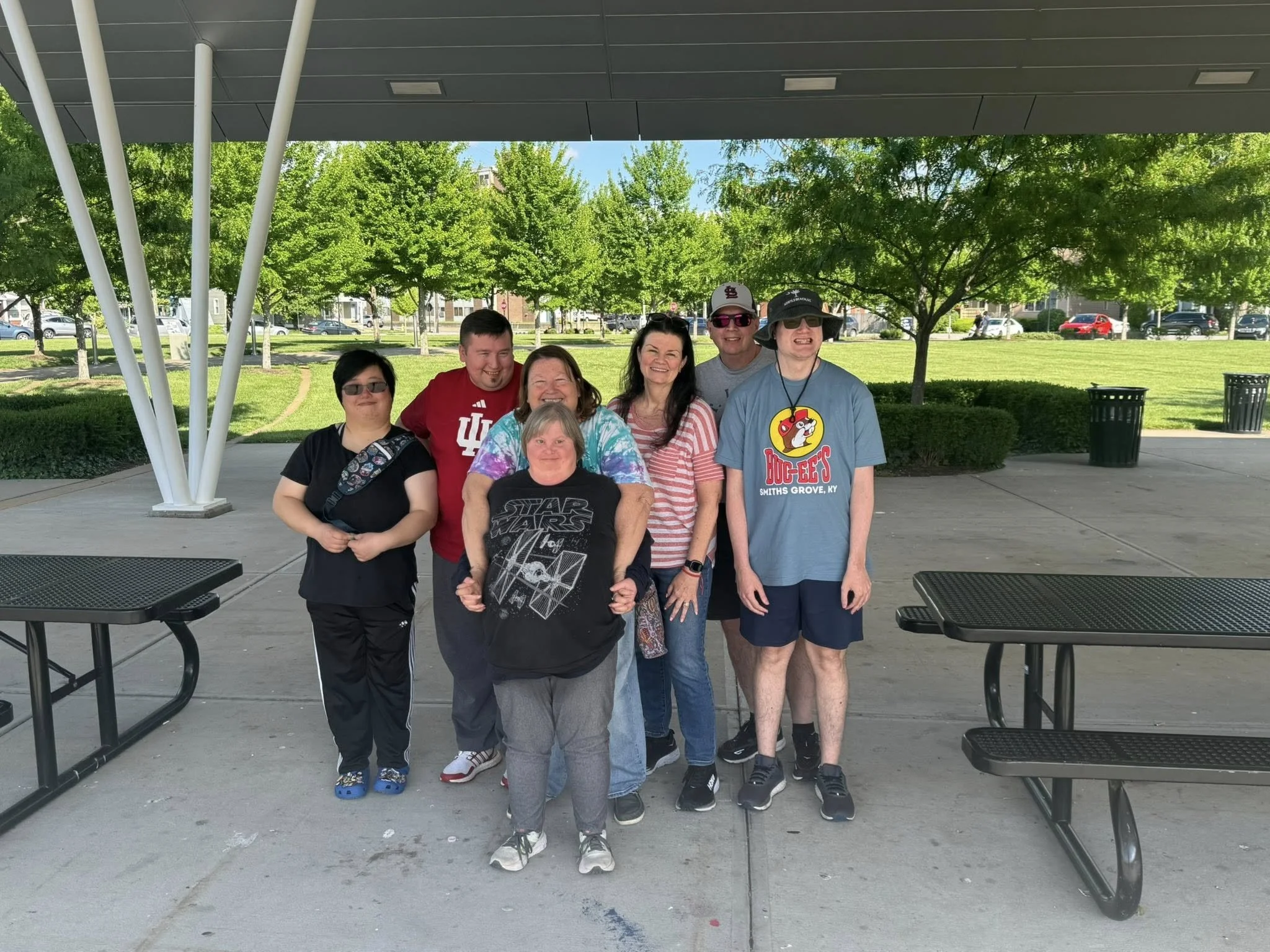 Group of seven smiling people standing outdoors under a pavilion with trees and parked cars in the background.