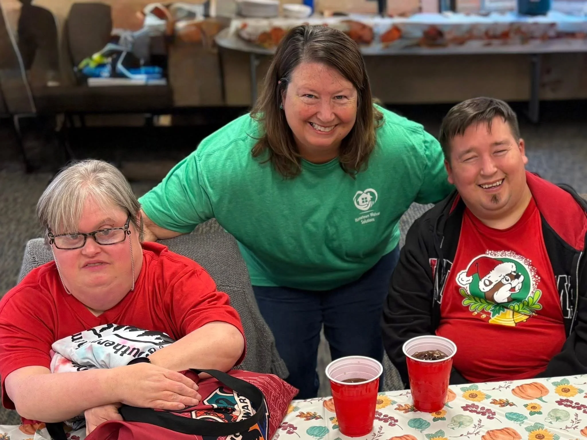 Three people smiling at a festive gathering, with two men seated at a table and a woman standing behind them, in front of a Thanksgiving-themed tablecloth and drinks.