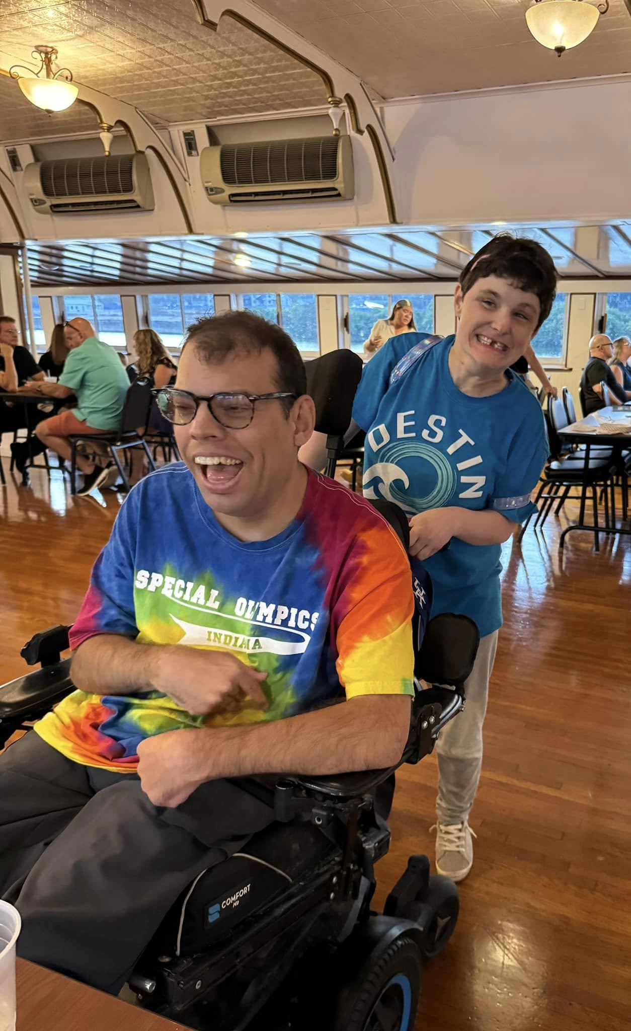 Two young men with disabilities enjoying time together in a dining hall or restaurant with large windows, wooden floors, and ceiling lights, one sitting in a wheelchair wearing a colorful tie-dye shirt, the other standing behind him wearing a blue T-