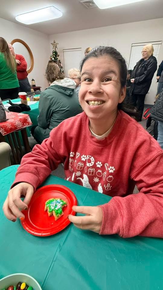 Young girl smiling and holding a decorated Christmas tree-shaped cookie on a red plate at a holiday gathering.