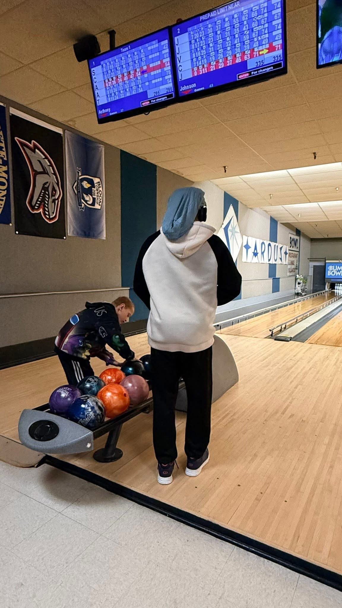 A boy selecting a bowling ball at a bowling alley, with a person standing nearby. The setting includes bowling lanes, a scoreboard overhead, and banners on the wall.