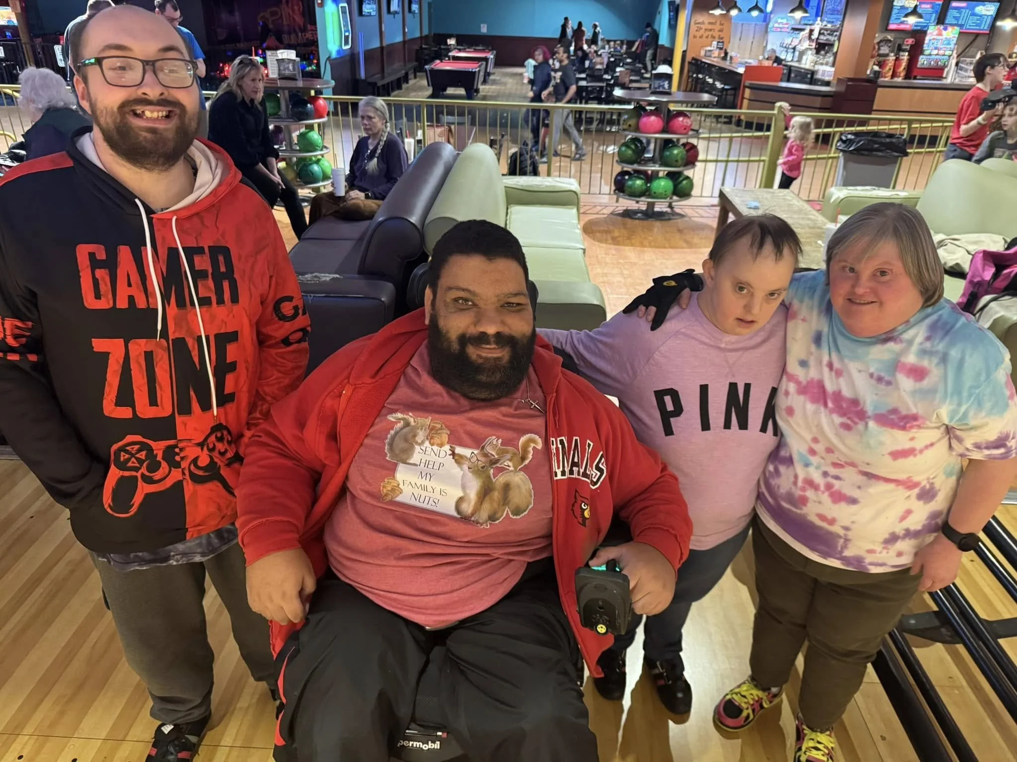 Group of four people at a bowling alley, smiling and posing for a photo. The background shows bowling lanes, balls, and other patrons.