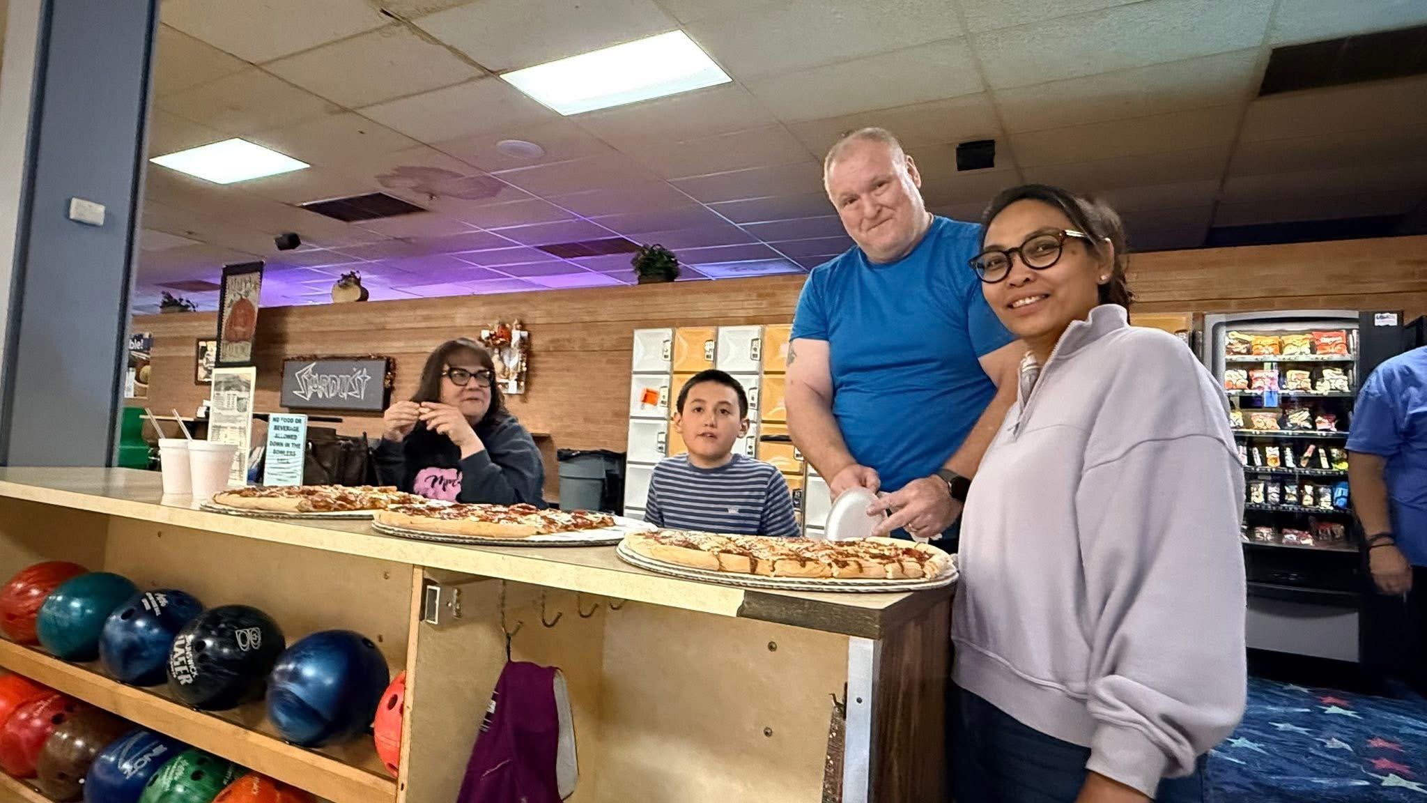A group of five people standing behind the counter in a bowling alley, with two pizzas and bowling balls in the foreground. They are smiling and posing for the photo.
