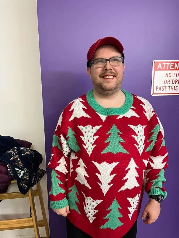 A man with glasses, a red cap, and a Christmas sweater with green and white Christmas trees, standing in front of a purple wall with a sign that says 'ATTEN...'