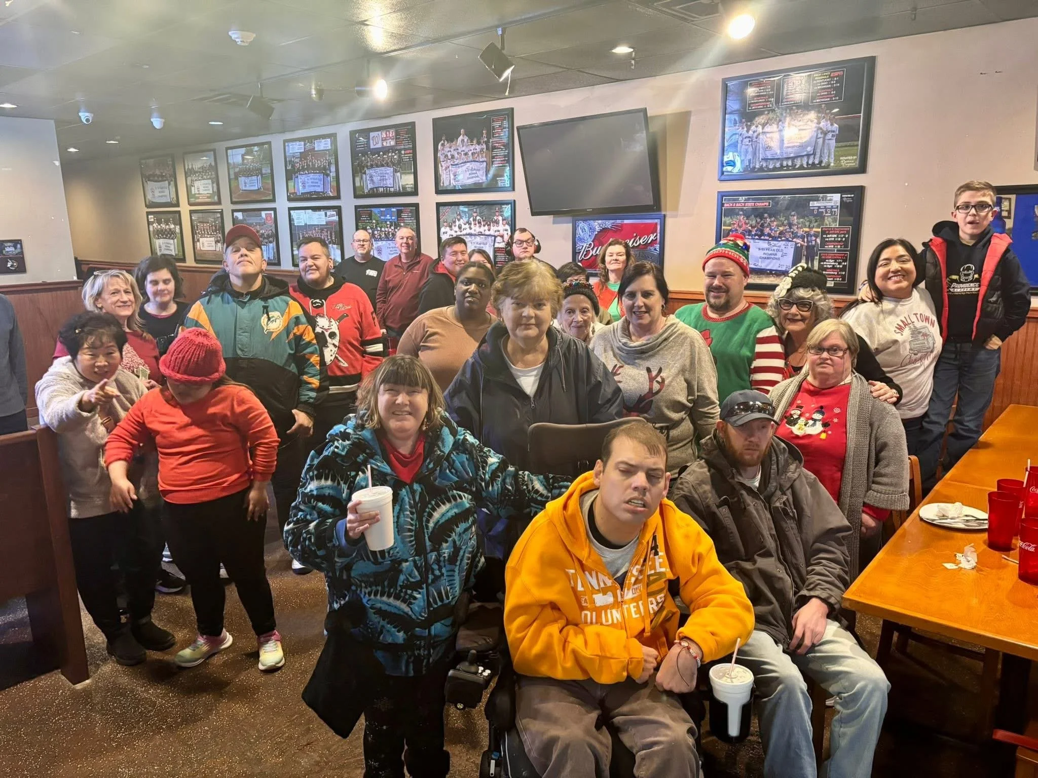 Group of people gathered in a restaurant or bar, some wearing holiday-themed sweaters and accessories, with multiple television screens on the wall behind them displaying sports games.