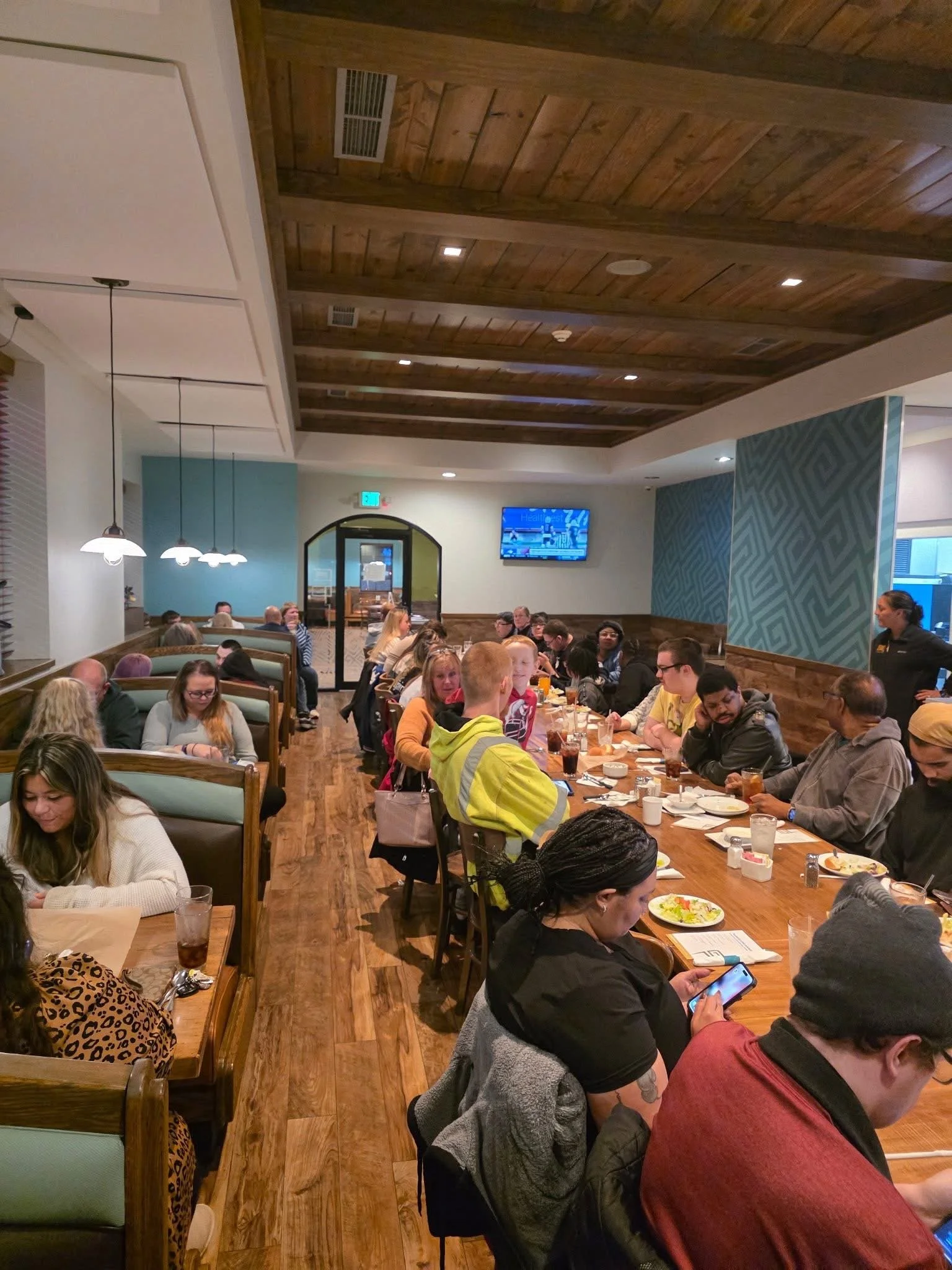 People dining in a restaurant with wooden ceiling and flooring, sitting at a long table, some using phones, some eating and drinking, with a TV on the wall and a woman standing nearby.