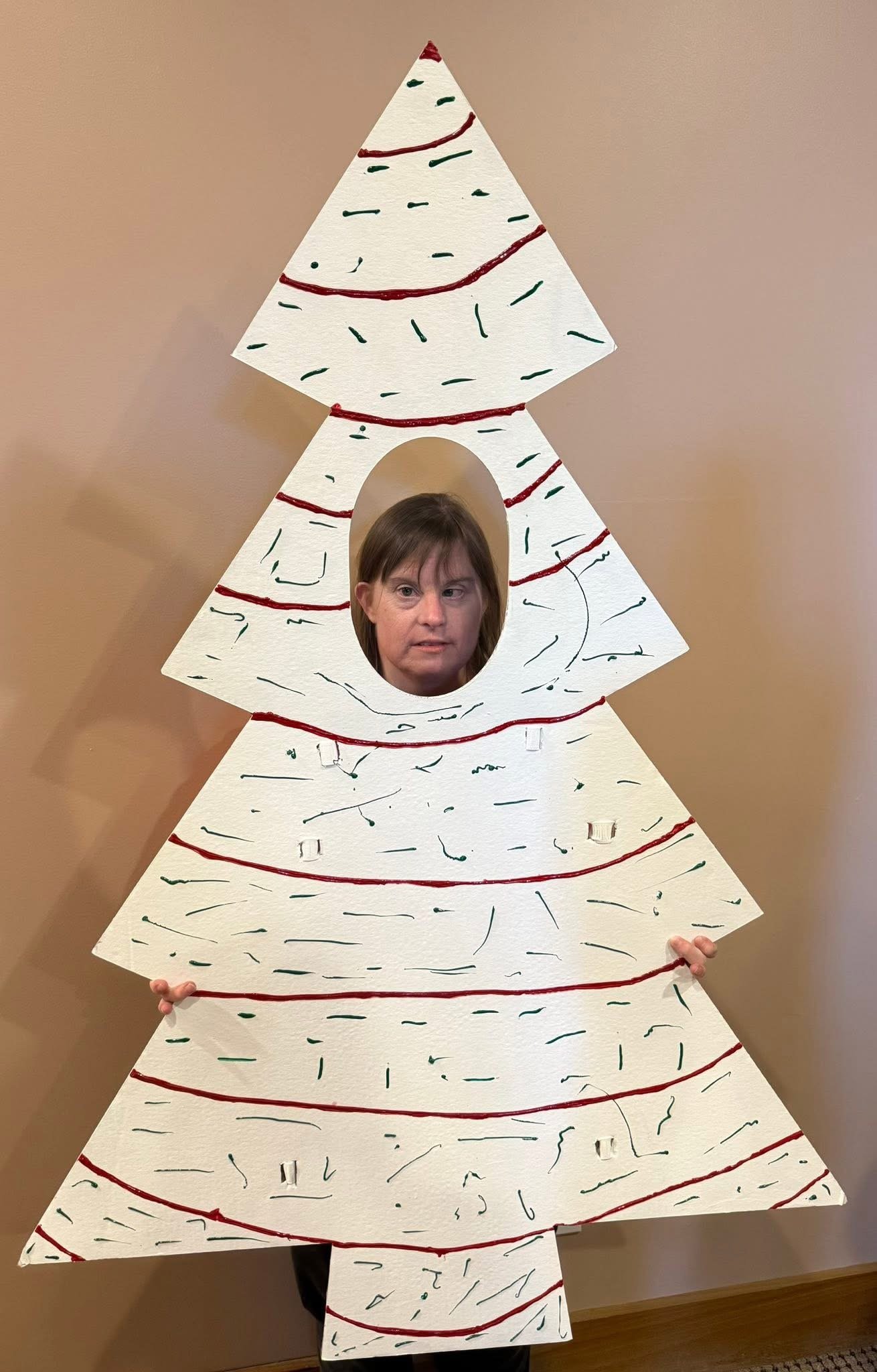 Person's head poking through a circular hole in a large, cardboard cutout of a decorated Christmas tree.