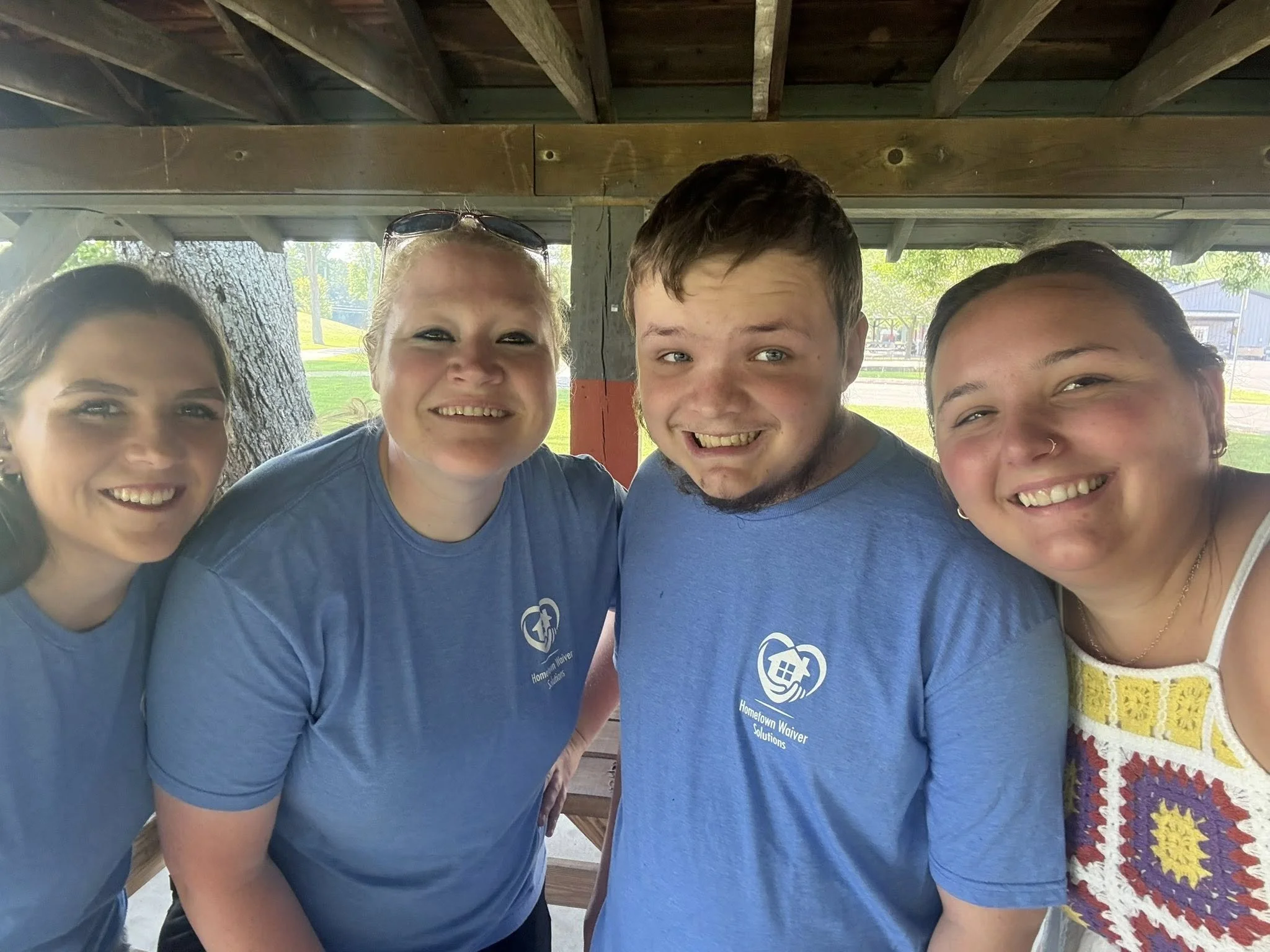 Four smiling people standing close together under a wooden pavilion in a park, with trees and a road in the background. Two women are wearing blue shirts with a logo that reads "Hometown Waiver Solutions."
