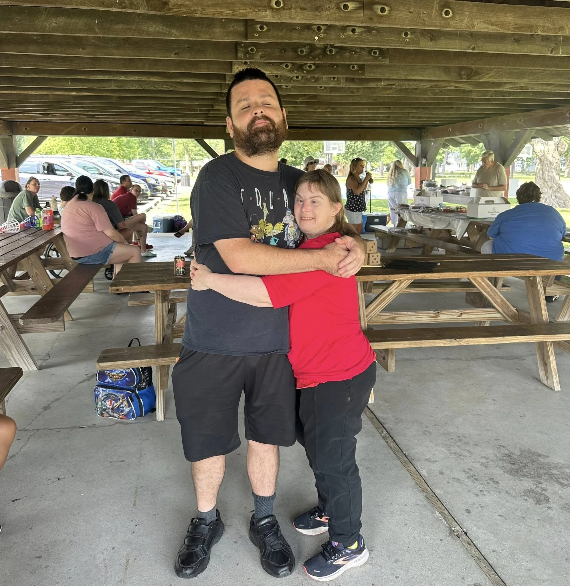 A man and woman hugging under a pavilion at a gathering, with picnic tables and other people in the background.