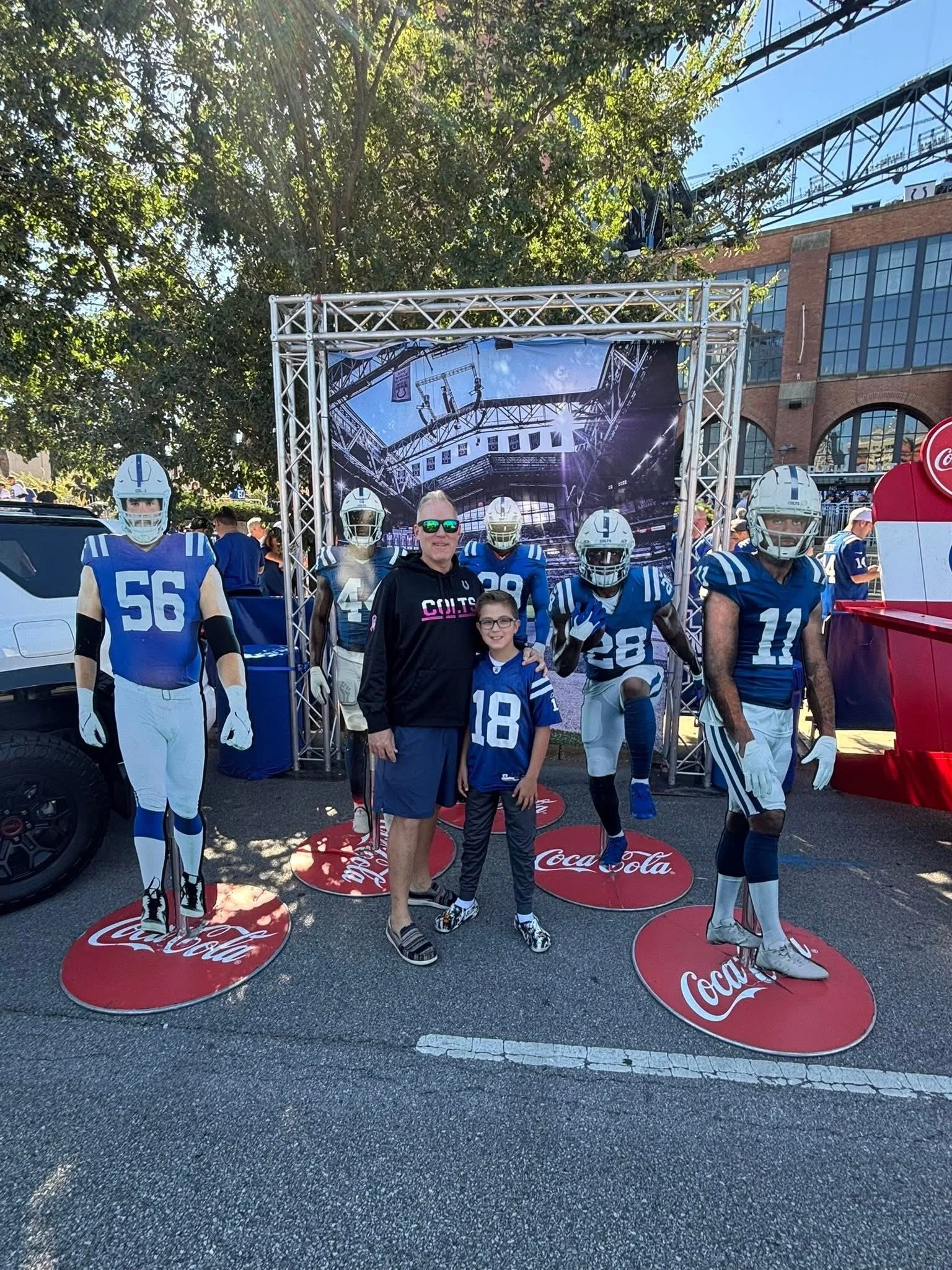 A man and a child standing together at a Coca-Cola display featuring mannequins in Indianapolis Colts football uniforms, with a background of trees and a building.